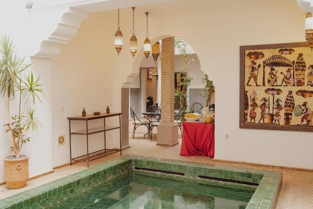 Courtyard with green pool, arched doorways, hanging lanterns, artwork, and a table with refreshments.