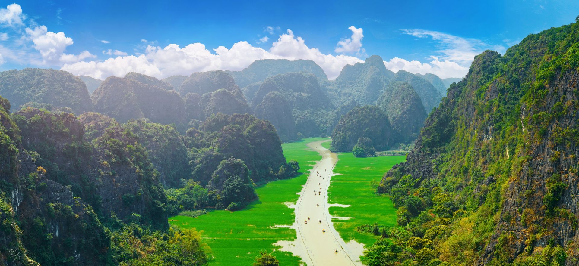 Mountains frame a river with boats, green fields, and blue sky.