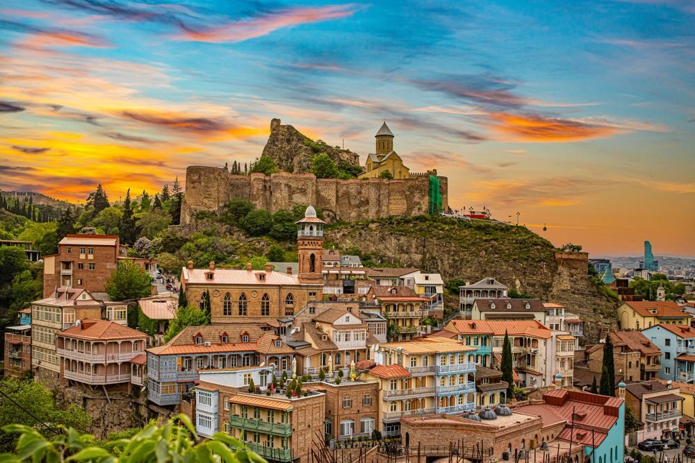 Cityscape of Tbilisi, Georgia, with old buildings, a fortress on a hill, and a colorful sunset sky.