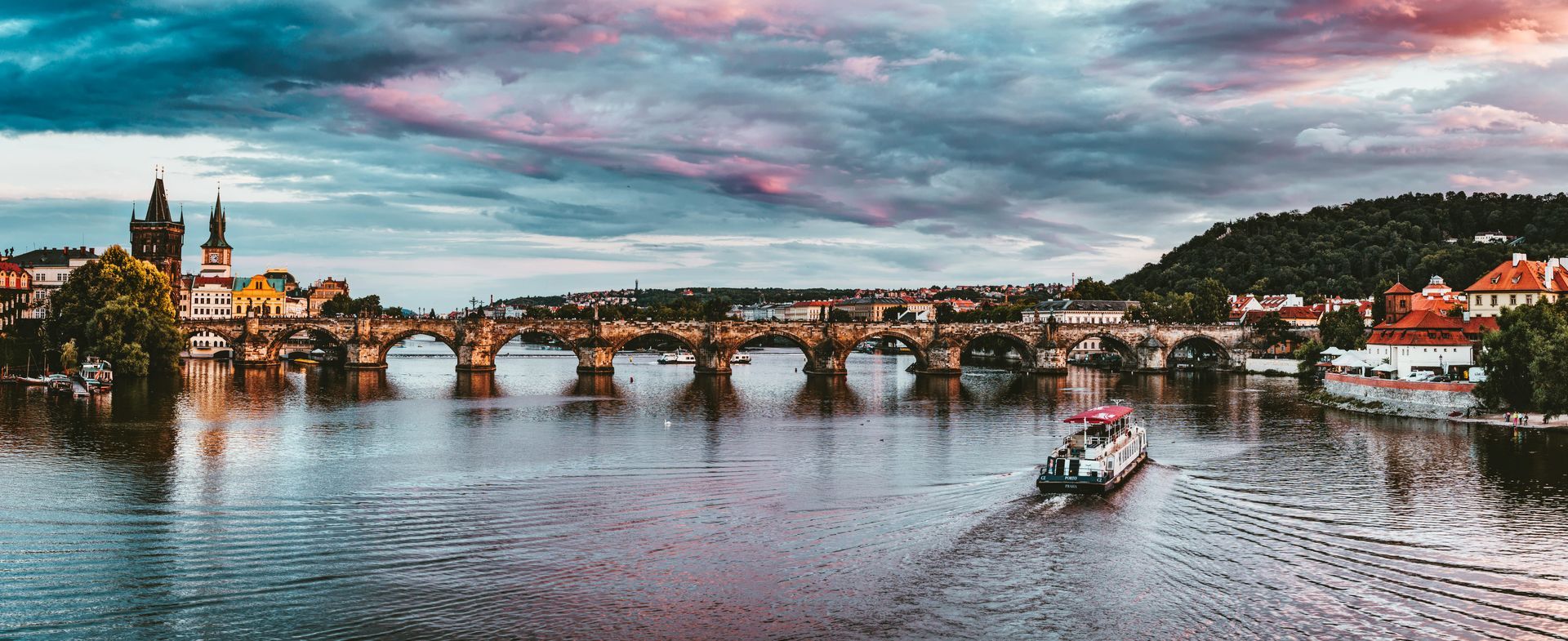 Prague cityscape with Charles Bridge and boat on Vltava River, under a colorful cloudy sky.