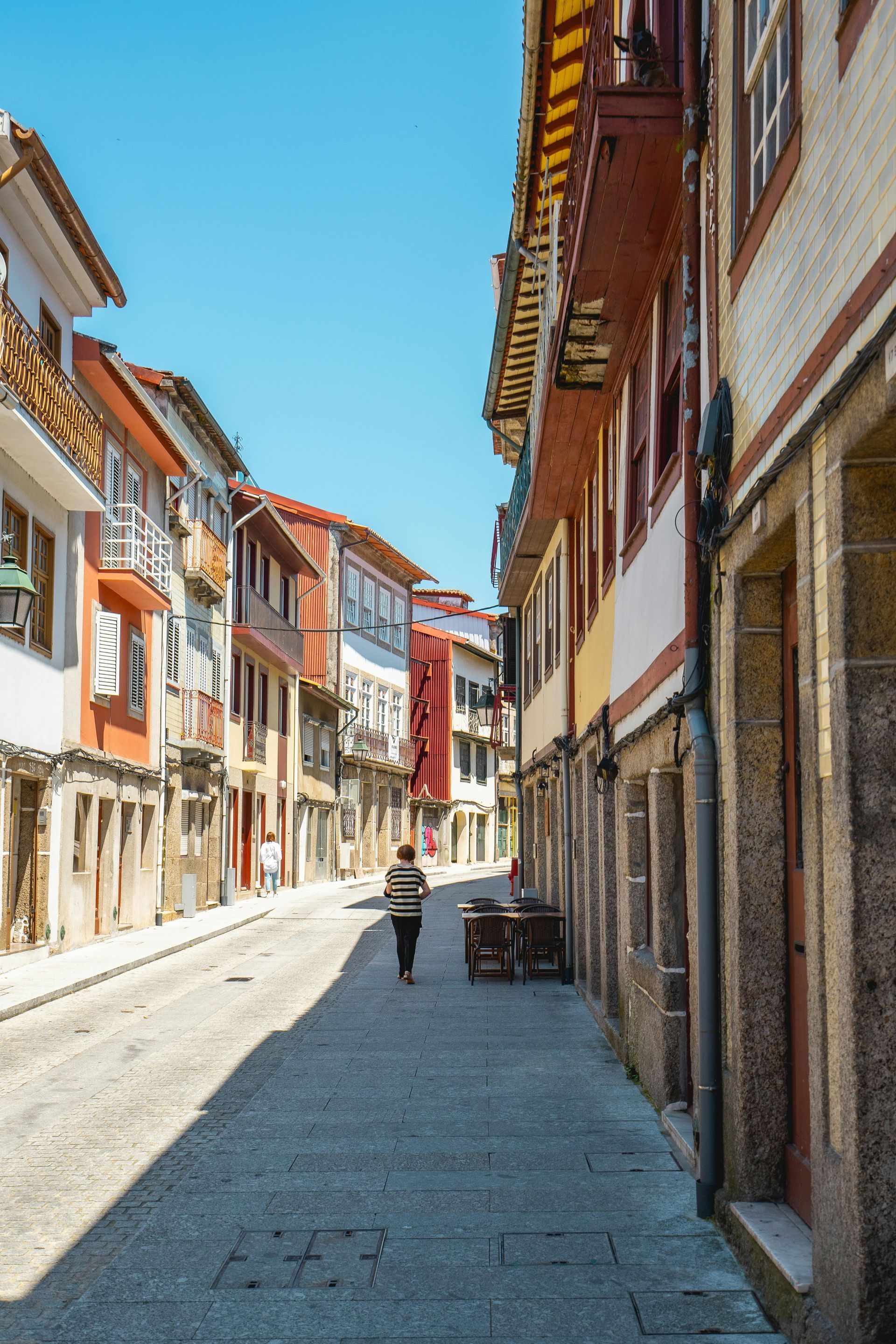 Narrow cobblestone street lined with colorful buildings under a bright blue sky. A person walks down the street.