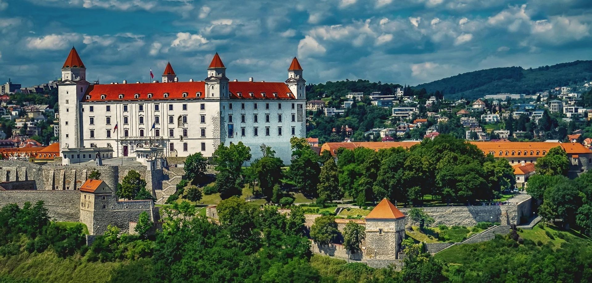 Bratislava Castle, a white rectangular building with red roofs, perched on a hill, overlooking a cityscape.