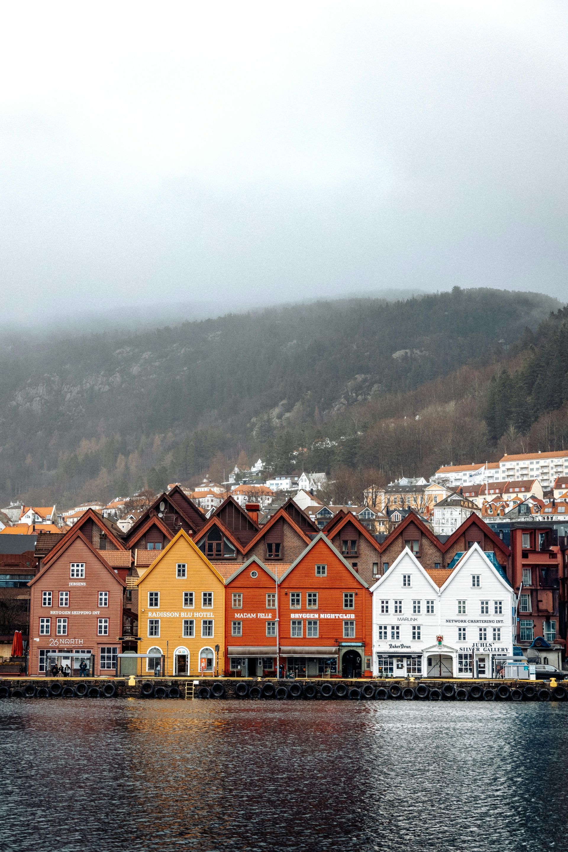 Colorful wooden buildings line a waterfront in Bergen, Norway, with a foggy mountain in the background.