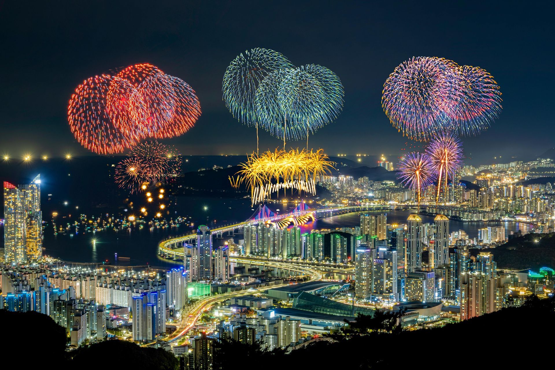 Fireworks over a cityscape at night; various colors illuminate the sky above the buildings.