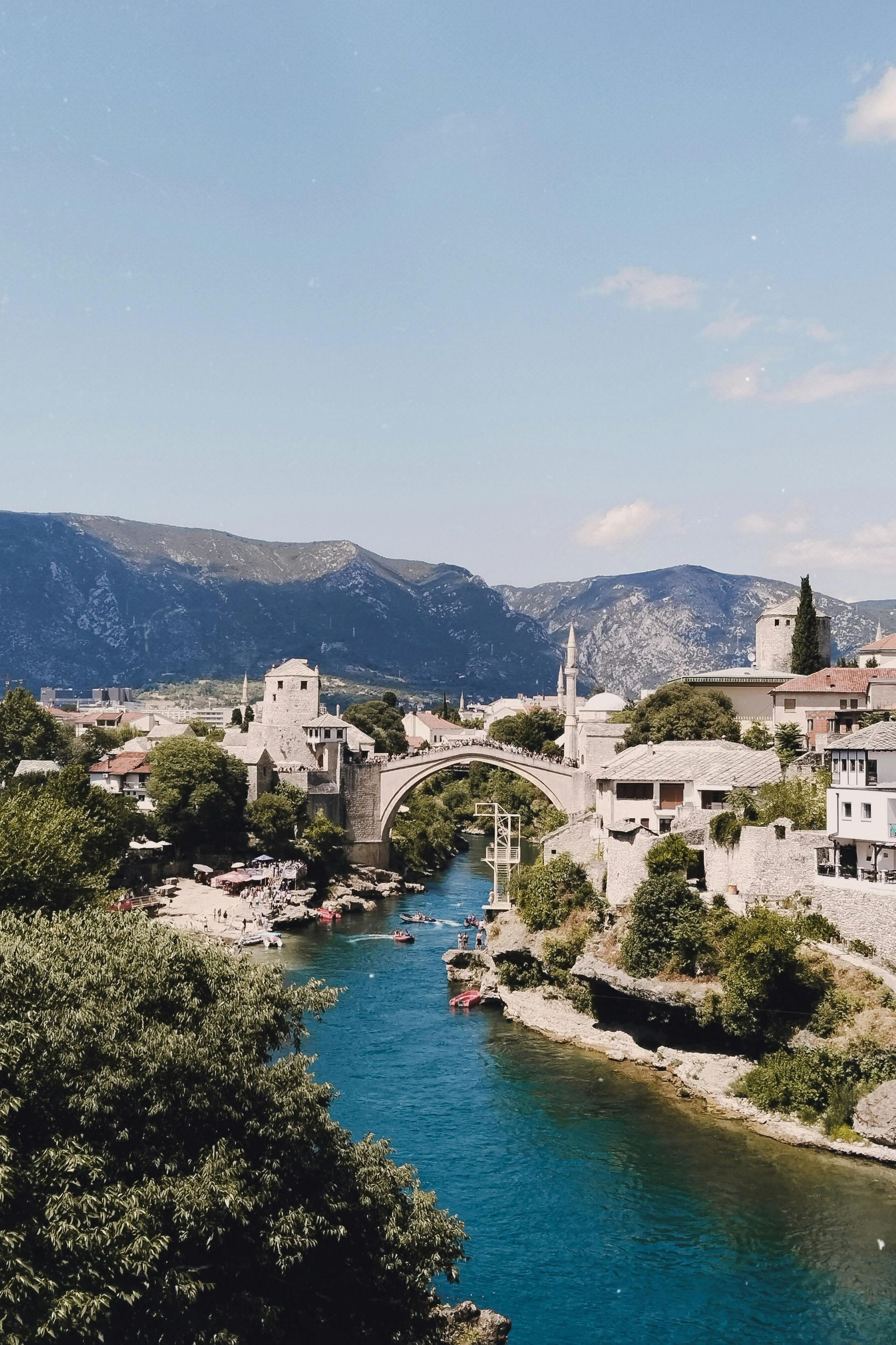 Old bridge over turquoise river in Mostar, Bosnia. Buildings line the riverbanks under a bright blue sky.