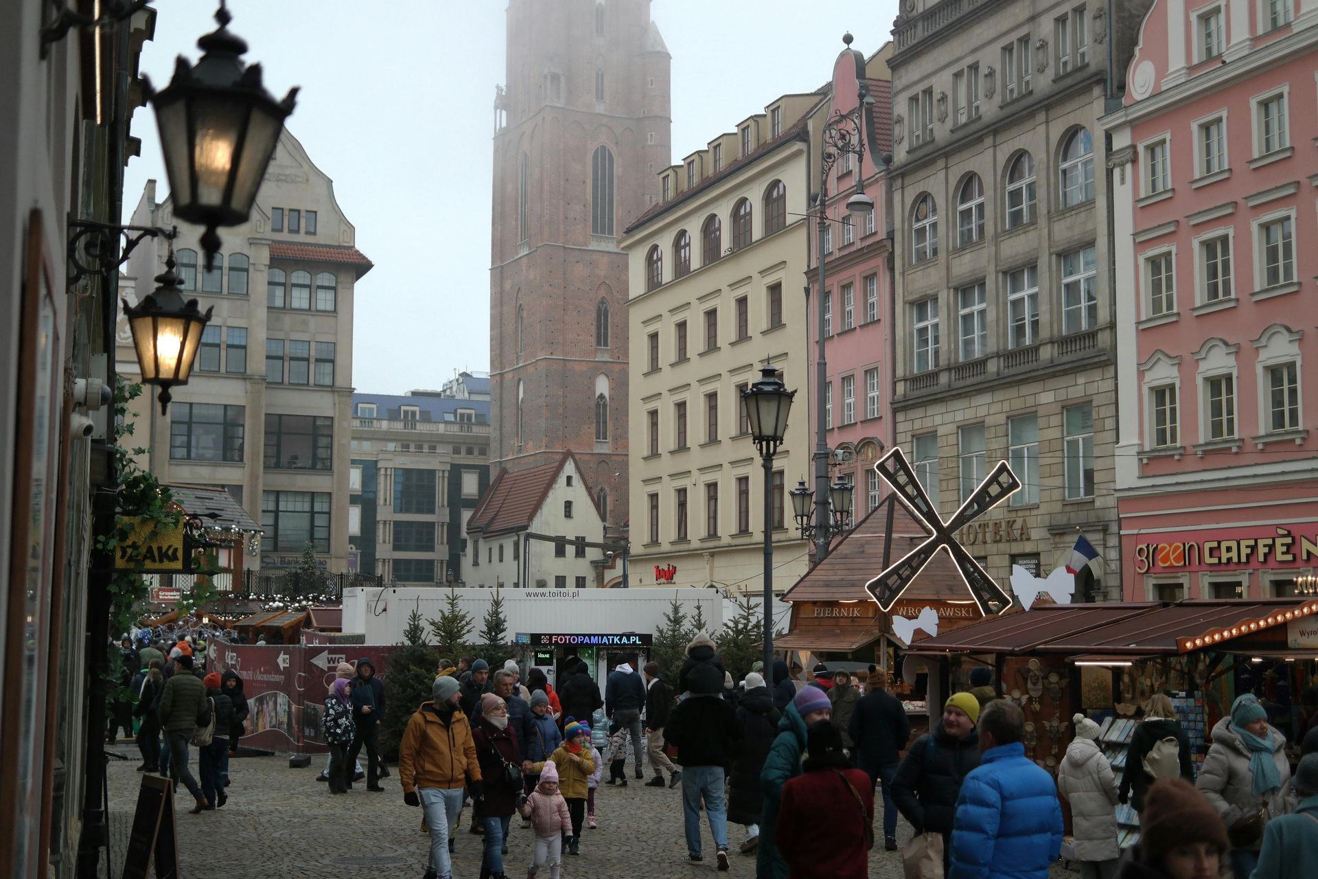Busy European square with Christmas market stalls, ornate buildings, and a tall church tower on a cloudy day.