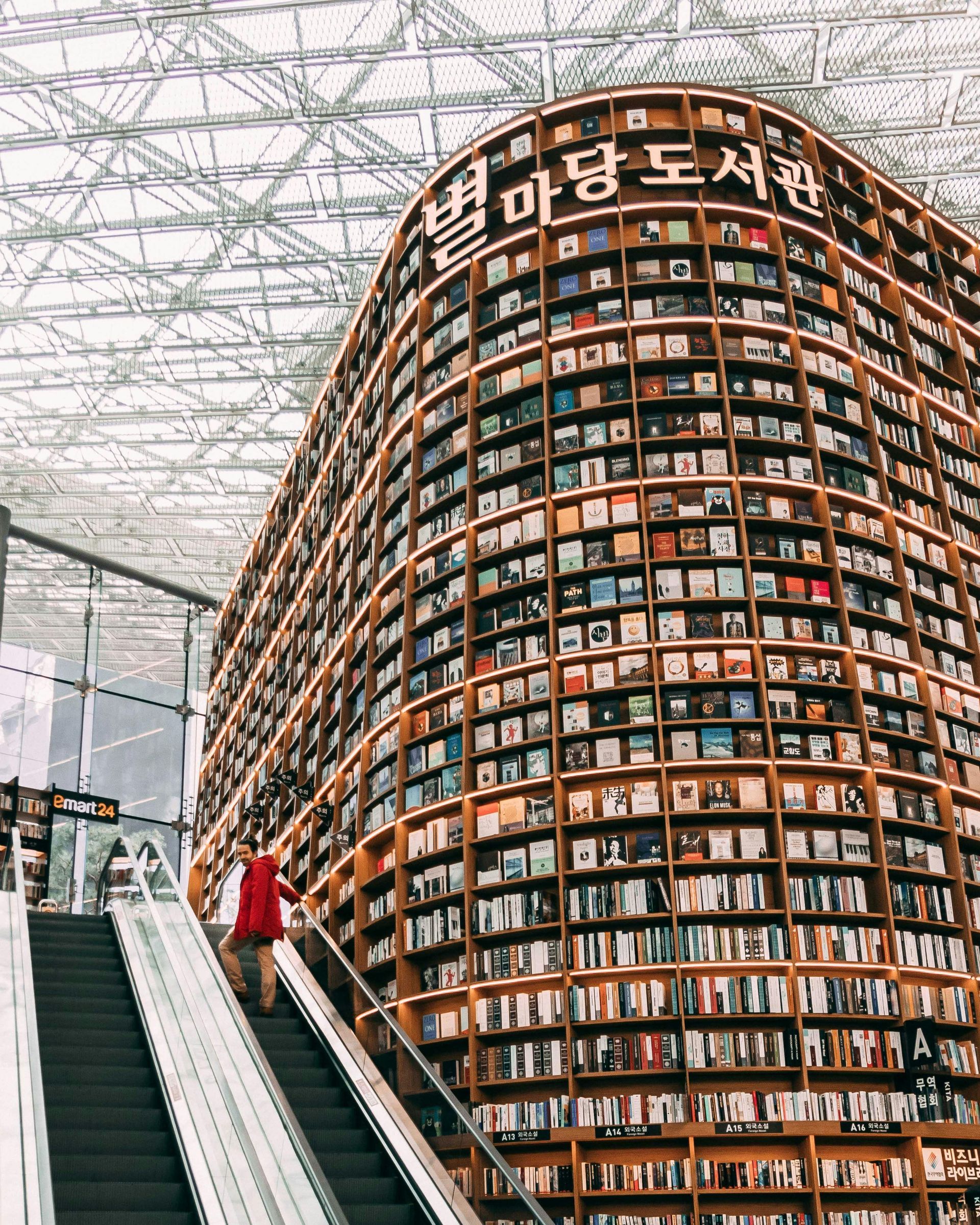 A person ascends an escalator, with a massive library of books stacked inside a building.