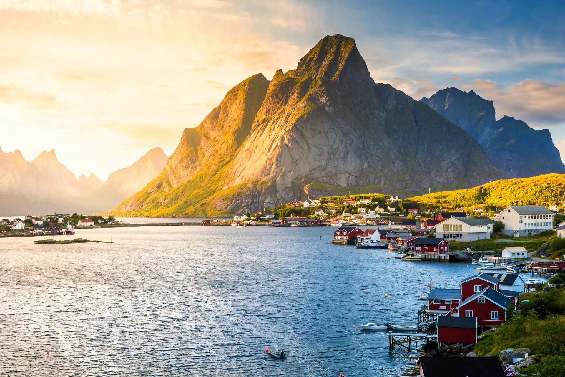 Coastal village with red and white buildings, mountains in the background, water in the foreground, sunny.