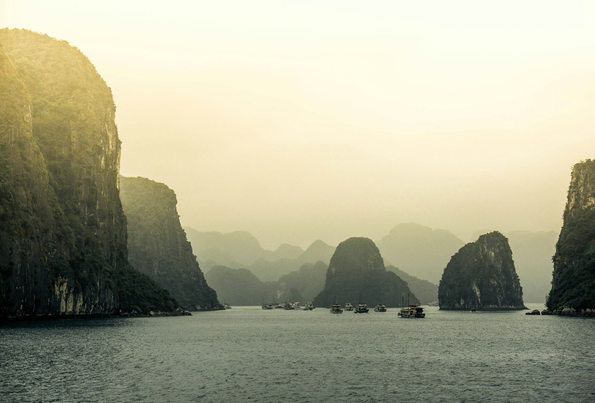 Limestone karsts in mist over water, boats visible. Soft yellow light.