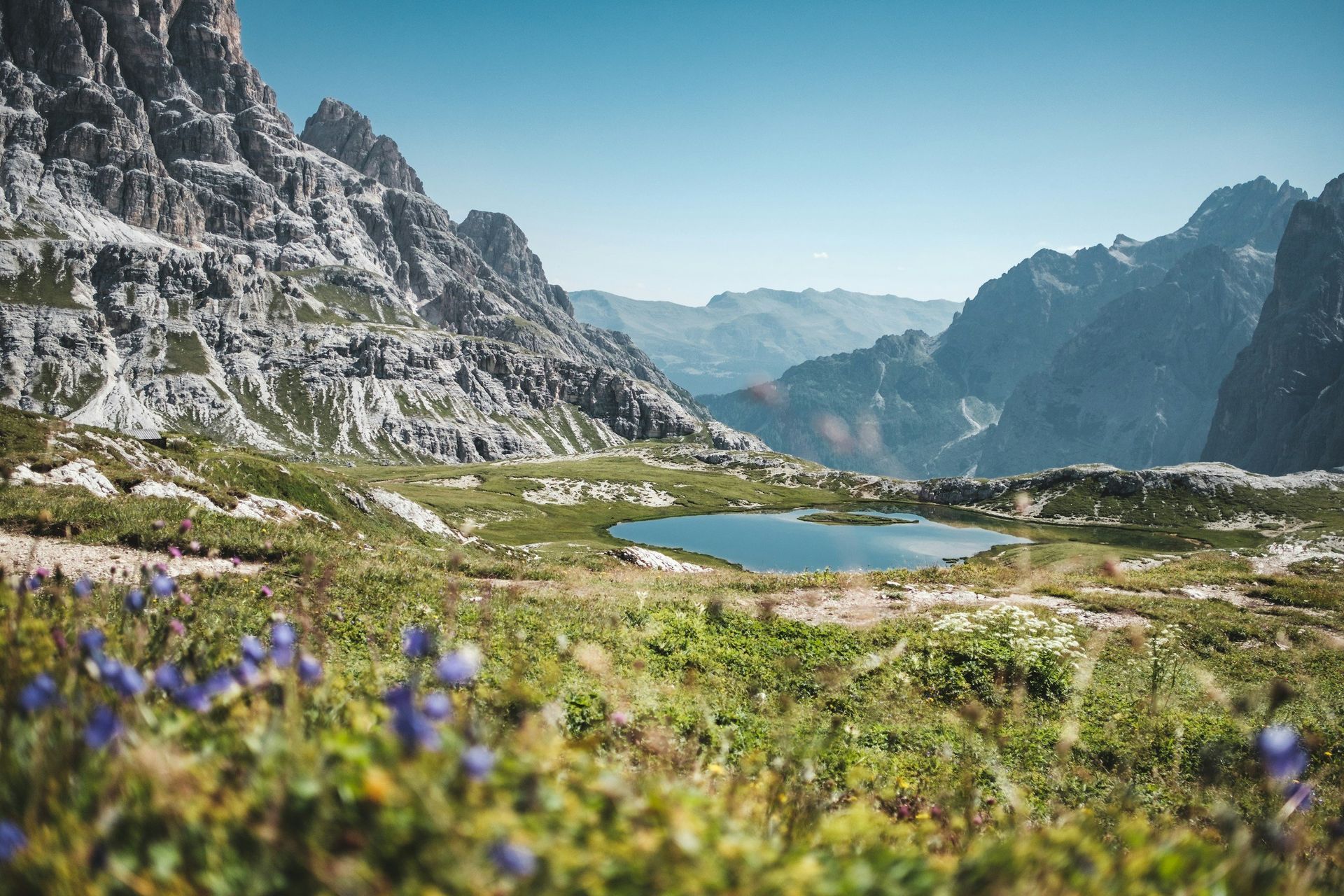 Mountain lake surrounded by rocky peaks and green fields under a clear blue sky.