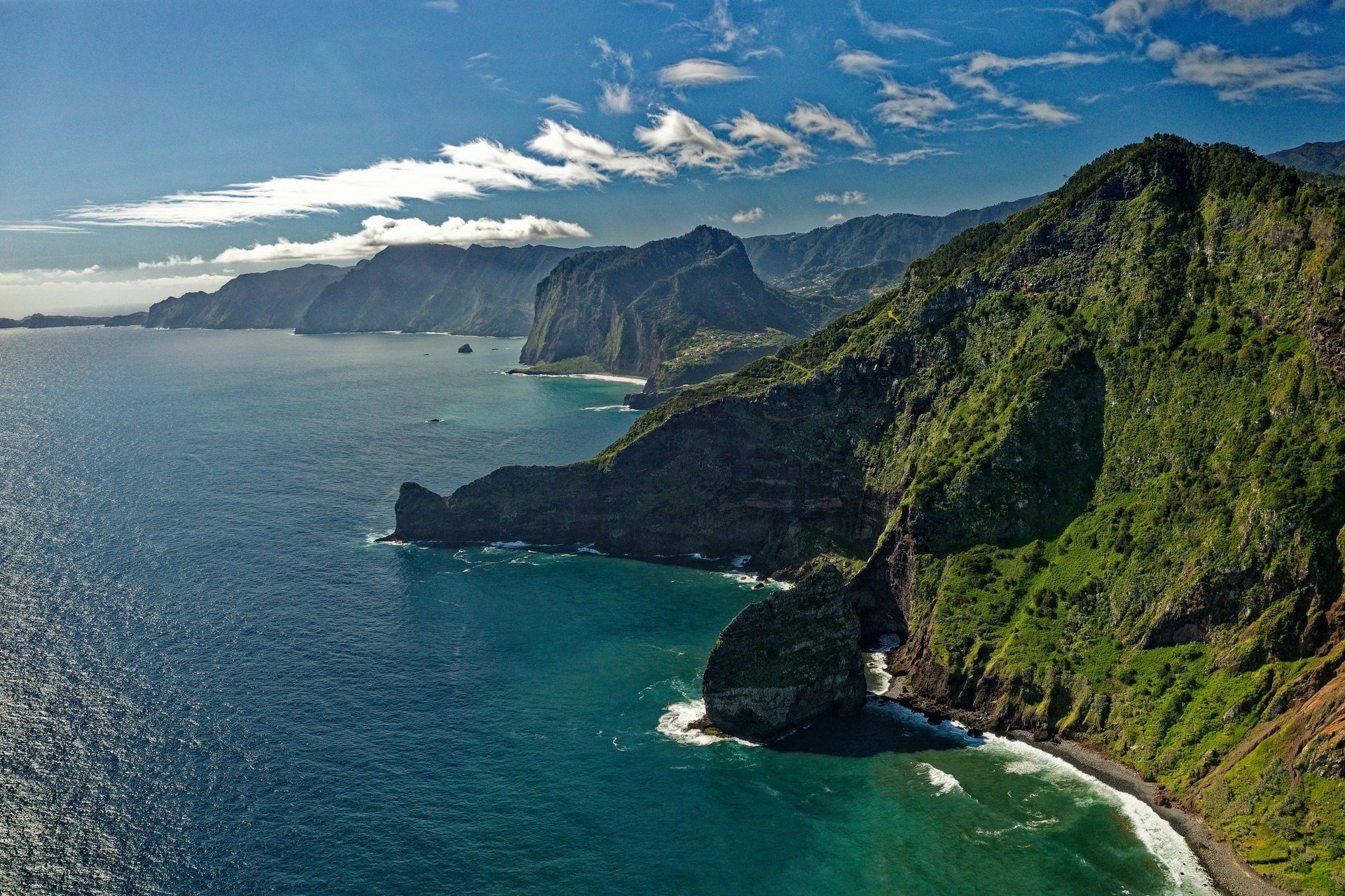Coastal view of steep, green cliffs meeting a deep blue ocean under a partly cloudy sky.