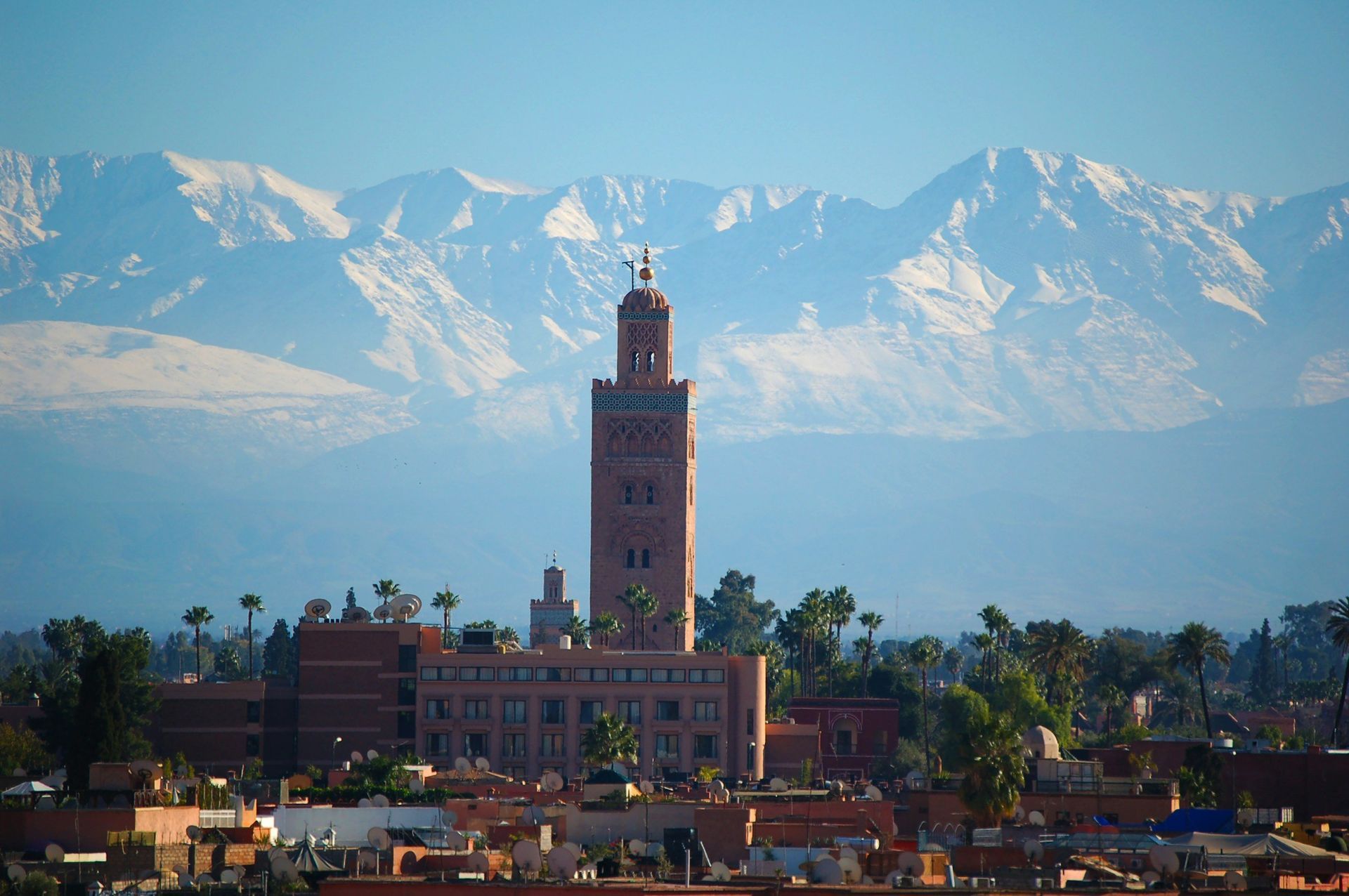 Koutoubia Mosque in Marrakech, Morocco, with snow-capped Atlas Mountains in the background under a blue sky.
