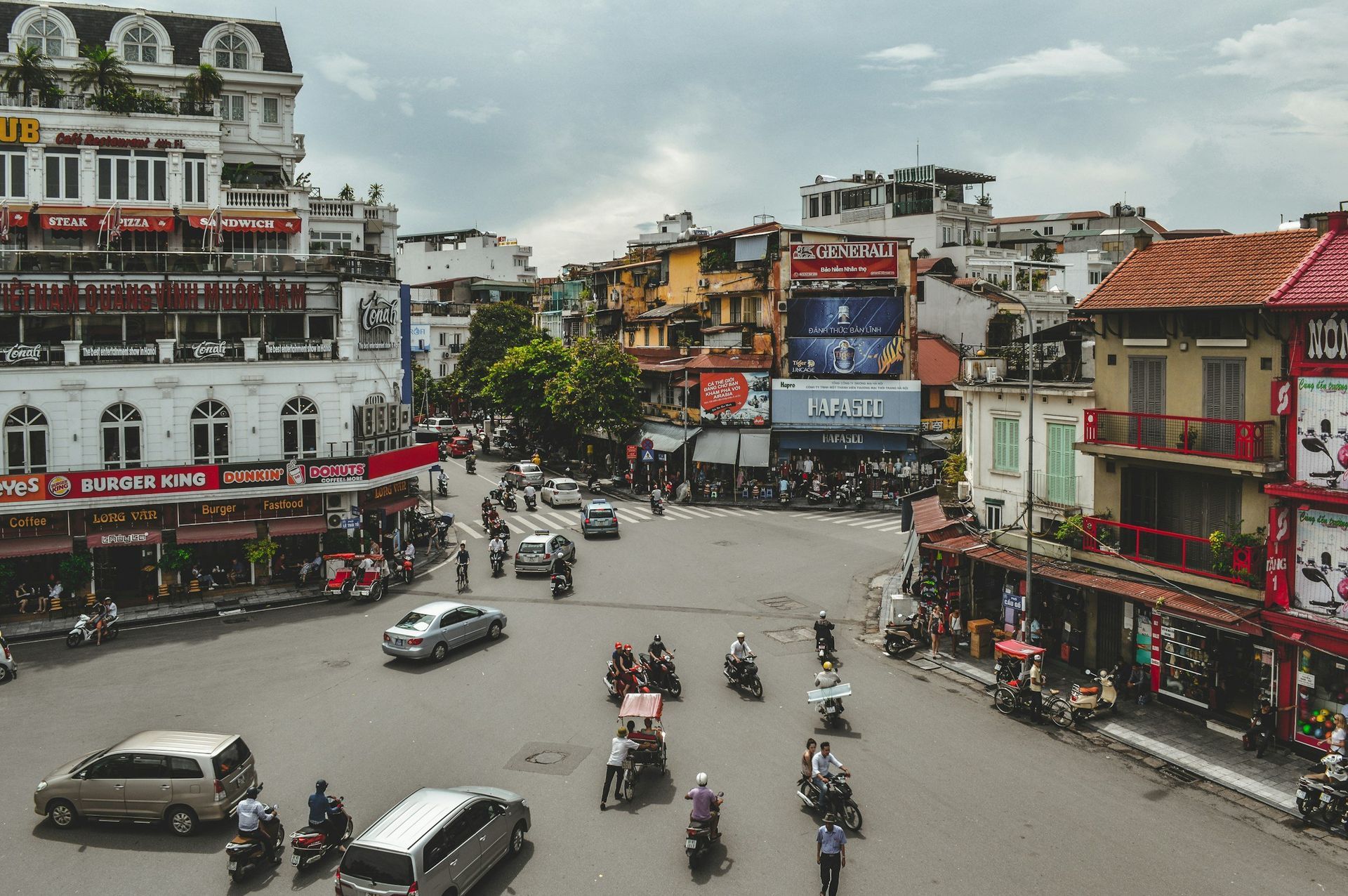 Busy city intersection with buildings, cars, and motorcycles; overcast sky.