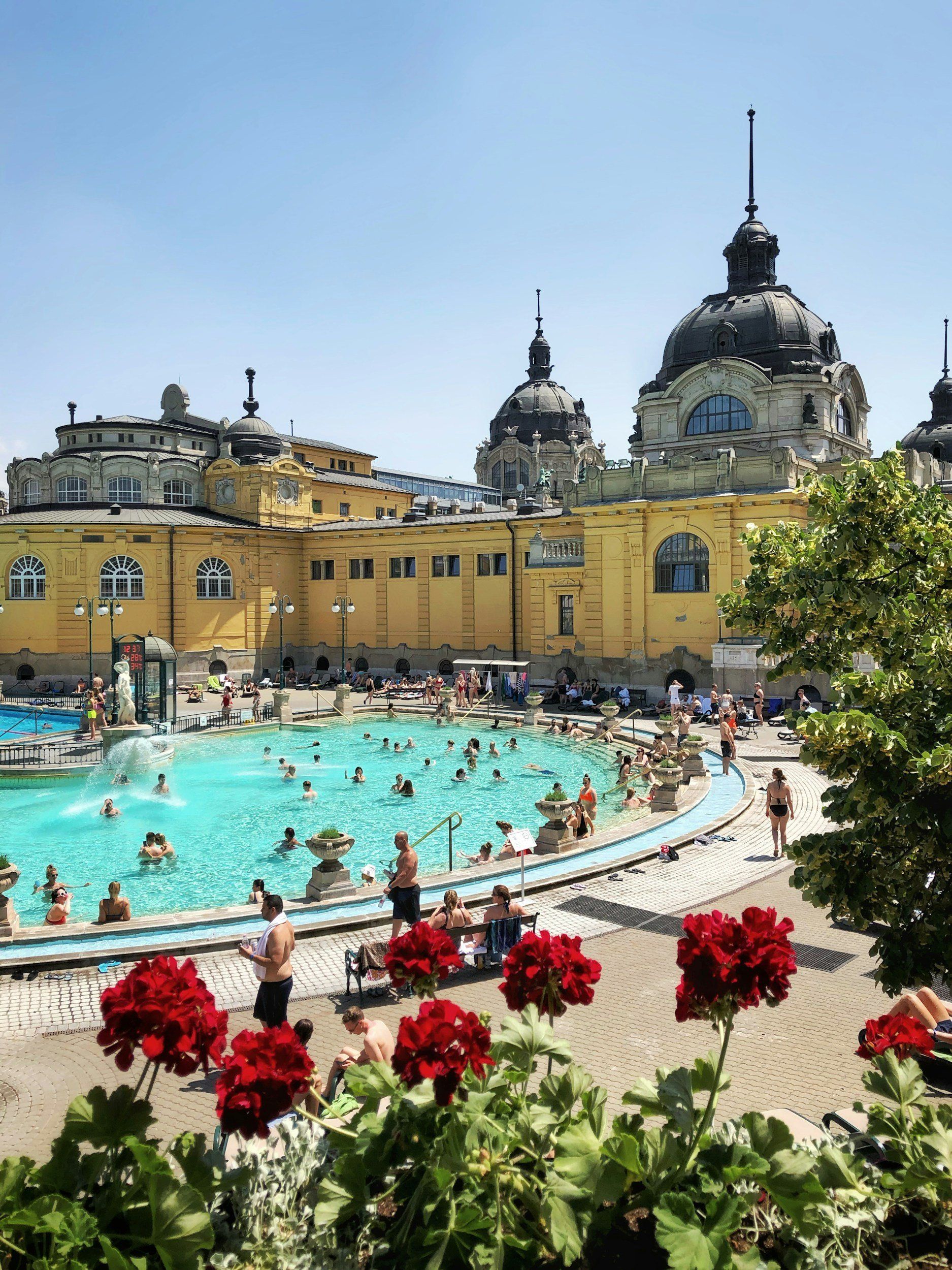 Outdoor thermal pool with people, historic yellow building background, red flowers in foreground.