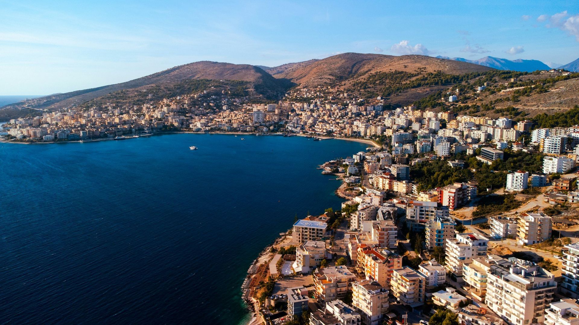 Coastal city nestled along a blue bay, with buildings and a mountainous backdrop under a clear sky.