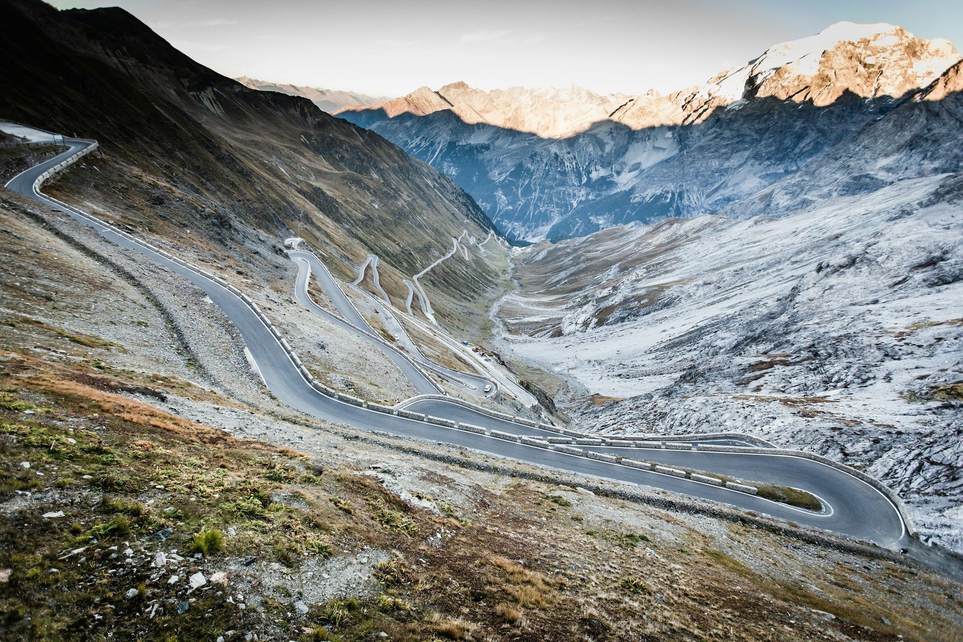 Winding mountain road with hairpin turns; landscape includes snow-covered peaks and a valley.