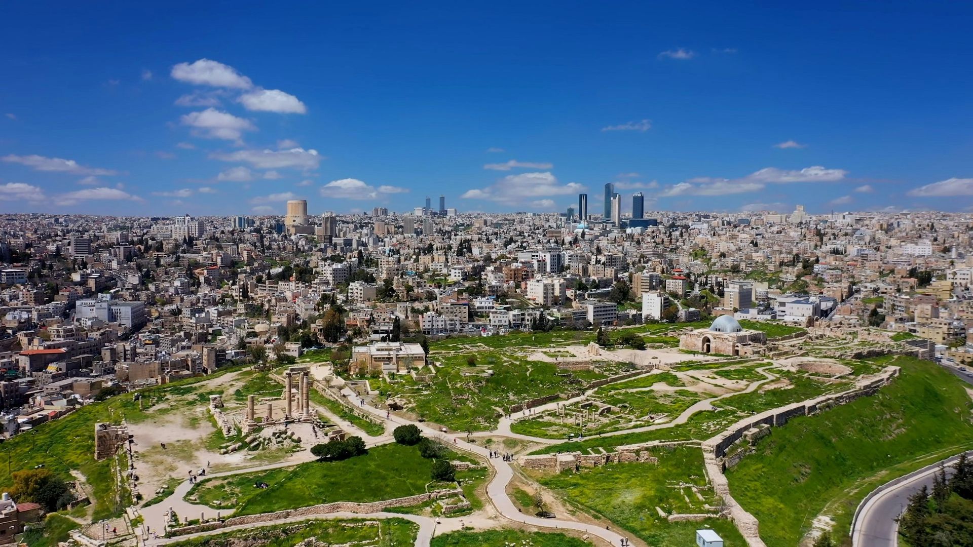 Aerial view of Amman, Jordan, showing city buildings and ruins on a hill under a blue sky.