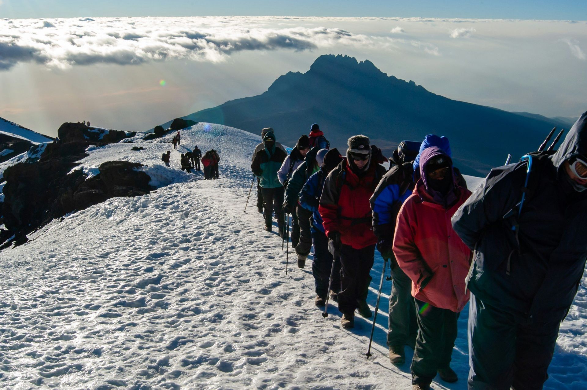 Hikers ascending a snow-covered mountain path towards a peak; cloudy sky in the background.