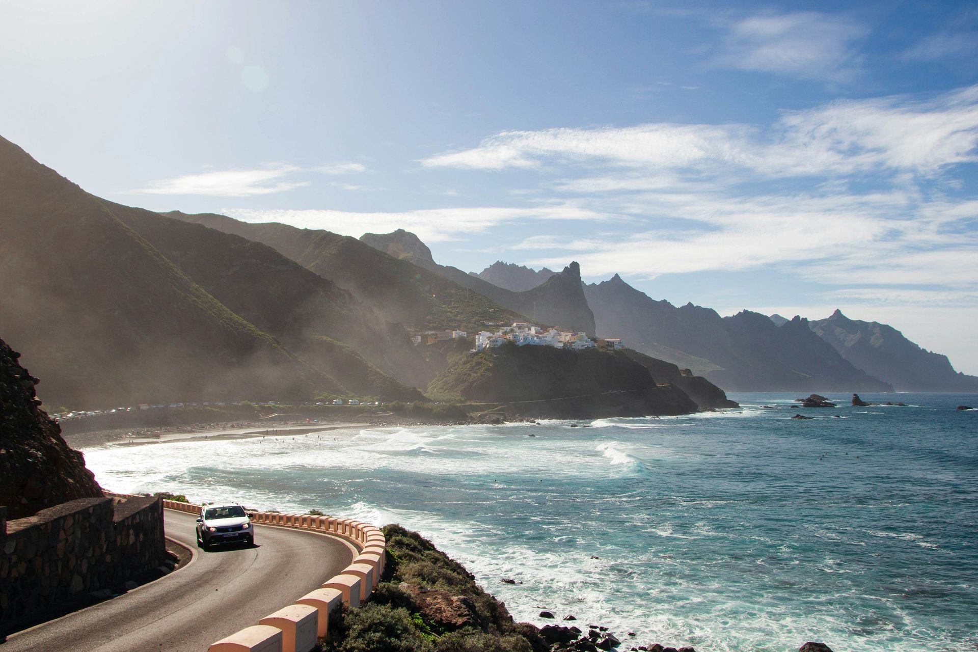 Car driving along a coastal road with ocean and mountain views under a blue sky.