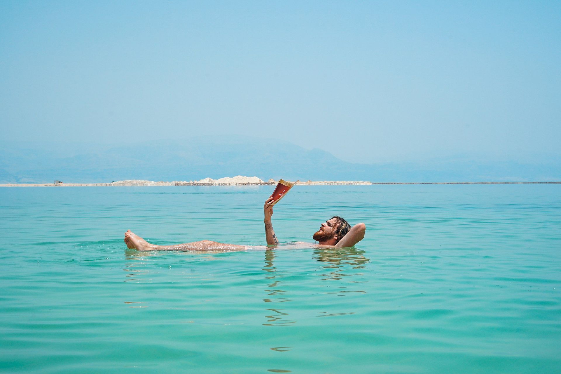 Man floating in clear water, reading a book, blue sky, mountains in the distance.