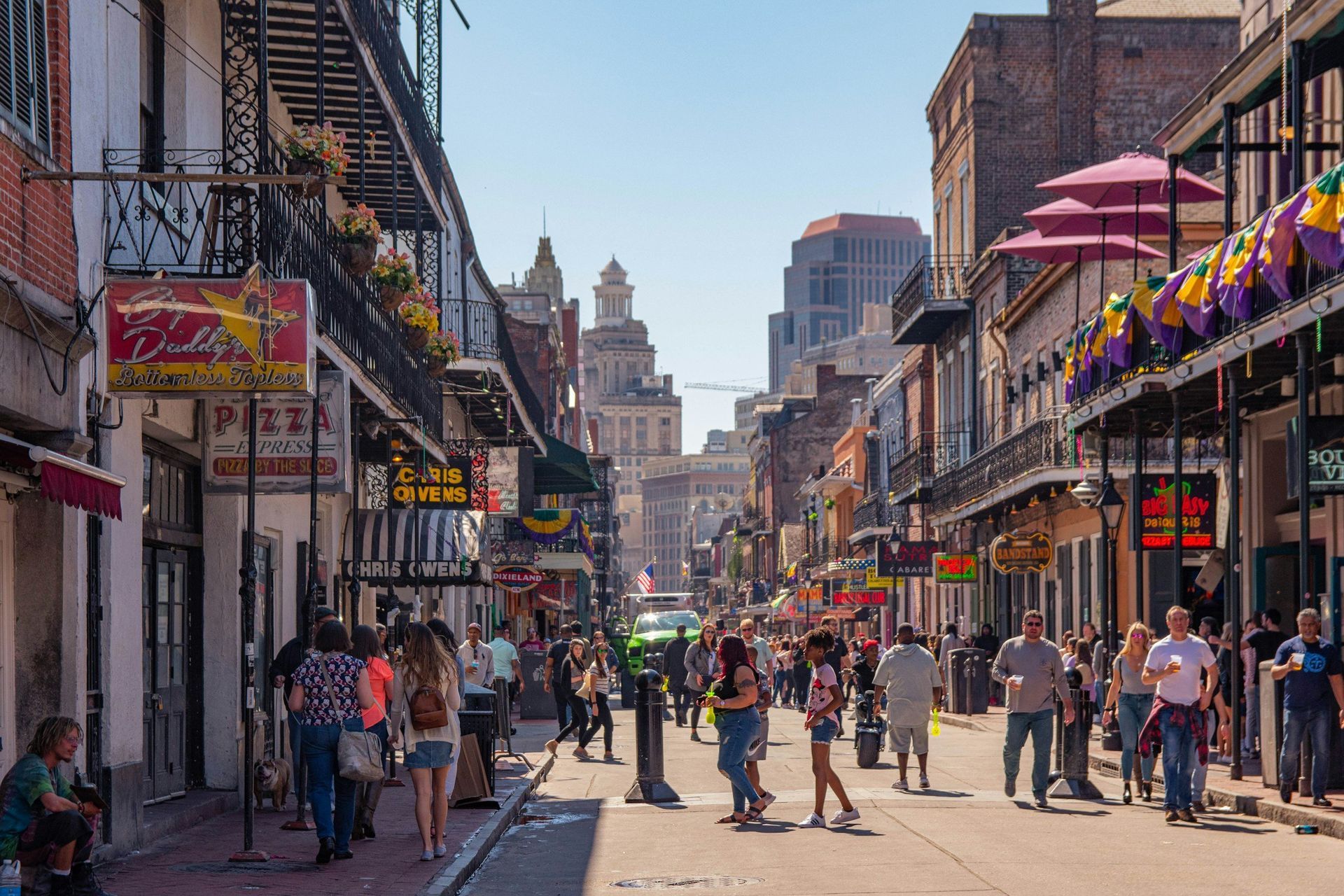 Street in New Orleans, lined with buildings, people walking. Sunny day, purple and yellow decorations.