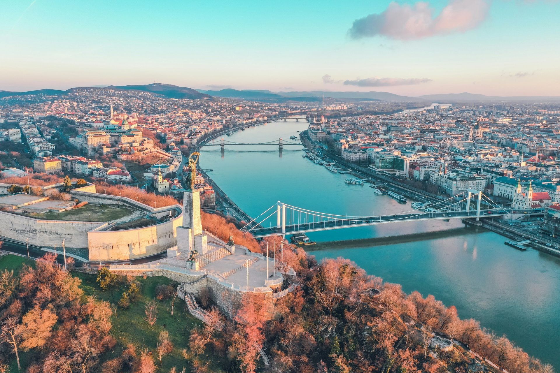 Aerial view of Budapest, Hungary, with the Danube River, bridges, and city buildings under a blue sky.