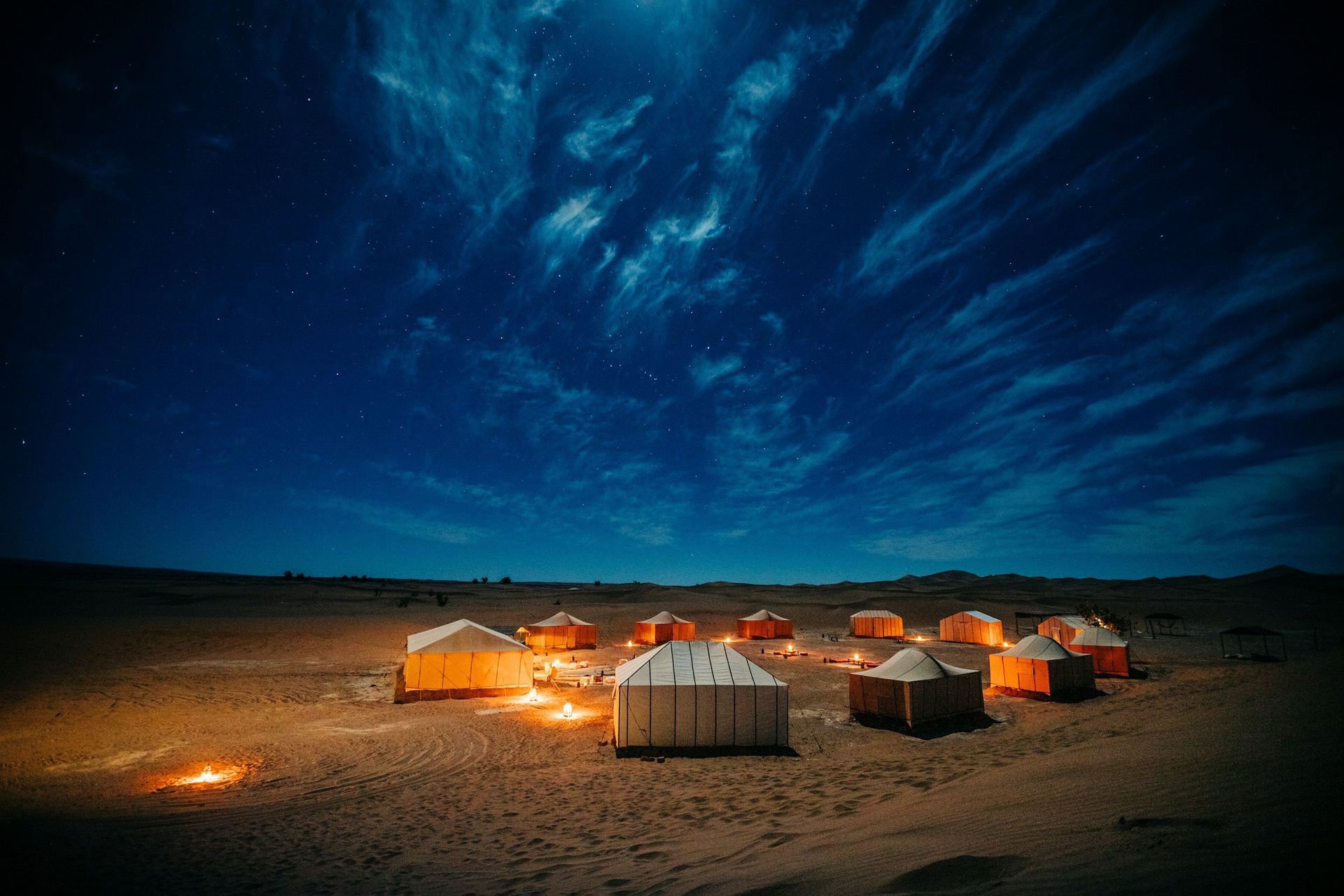 Desert campsite at night under a starry blue sky, with lit tents and fires.