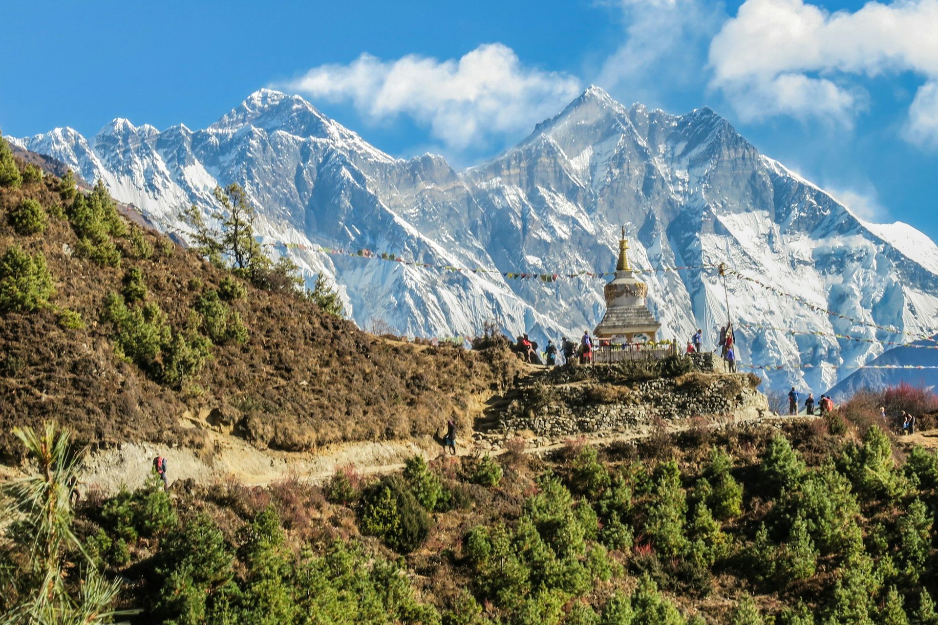 Snowy mountains behind a stone monument with people. Green hillside in foreground.