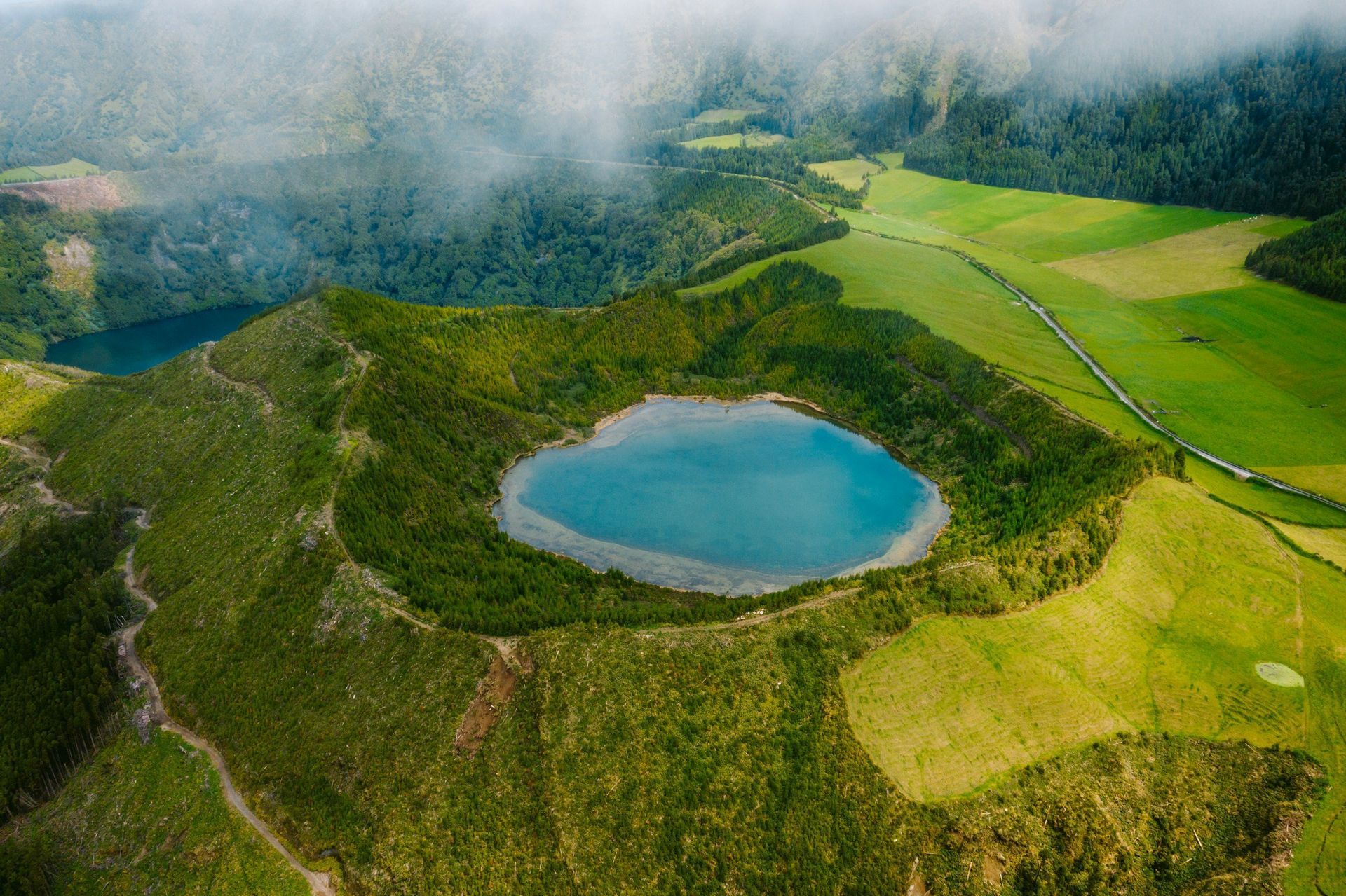 Aerial view of a heart-shaped lake in a volcanic crater, surrounded by green vegetation and a field, with fog in the background.