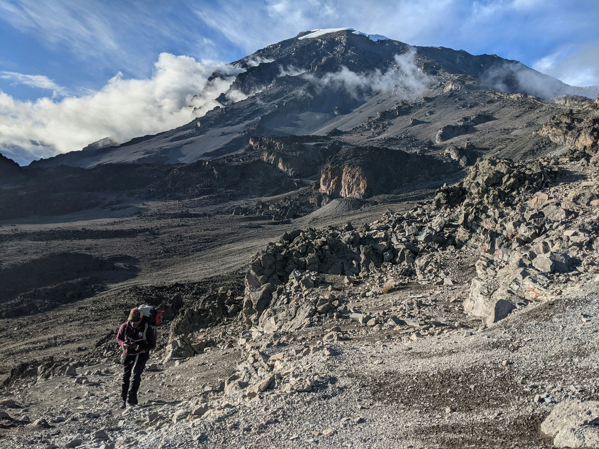 Person hikes on rocky terrain toward a snow-capped mountain.