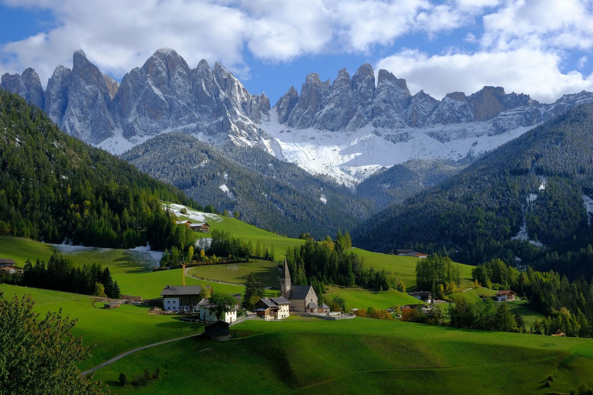 Green valley with small buildings and a church, snow-capped mountains in the background, blue sky.