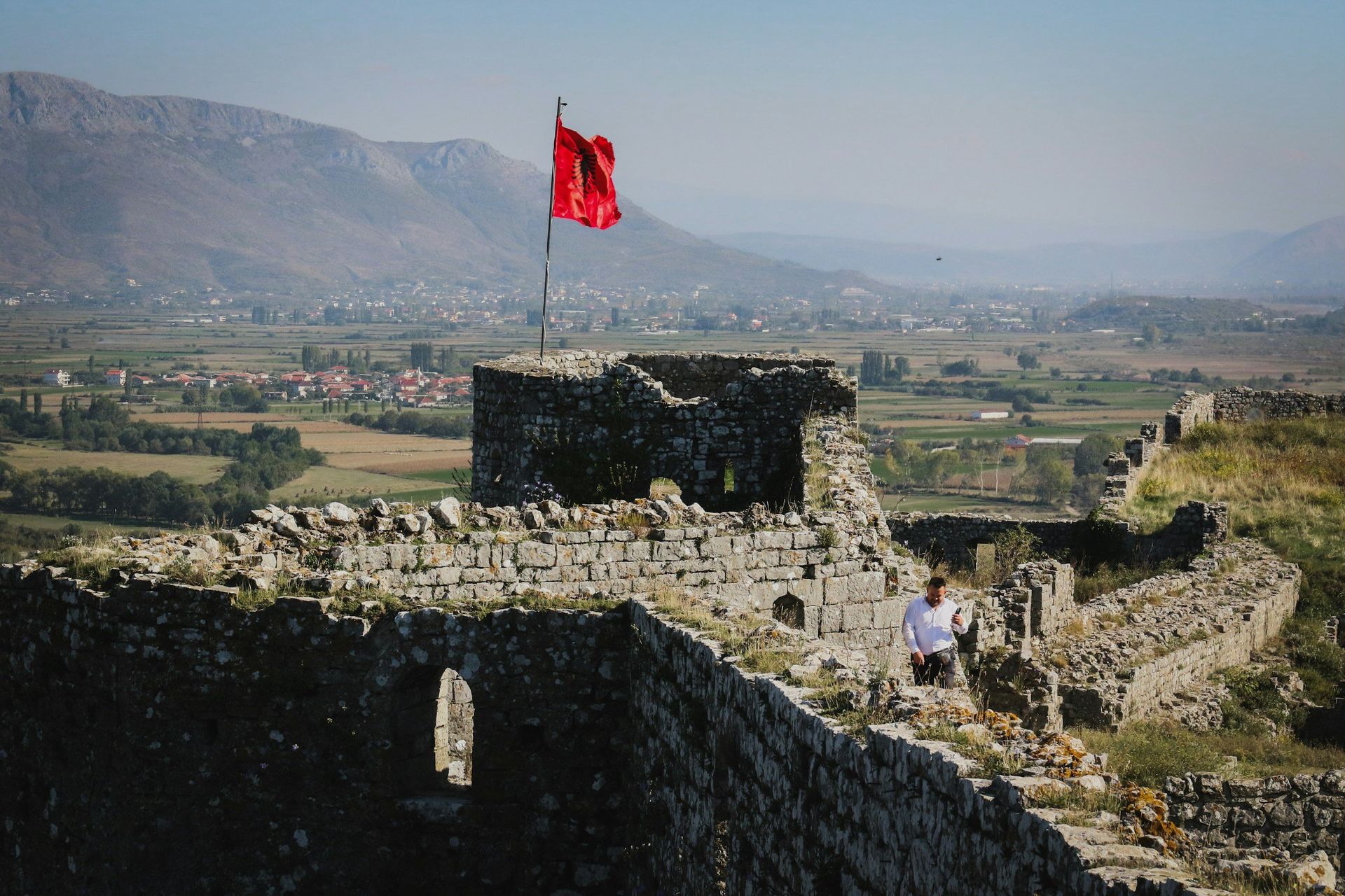 Ruin of Rozafa Castle with red flag waving; person walks atop stone wall with valley background.