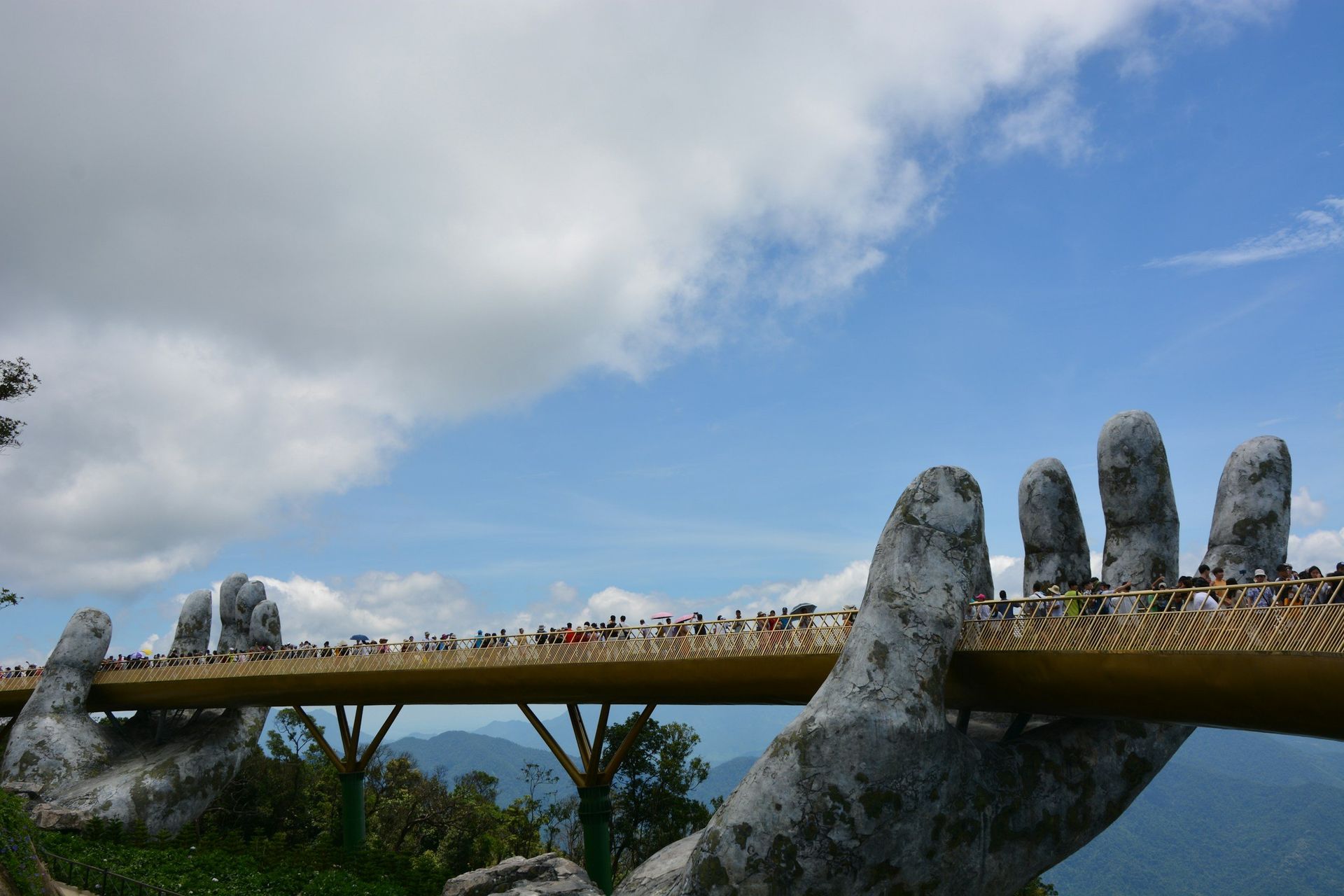 Golden Bridge held by giant stone hands, with people walking on it, under a blue sky.