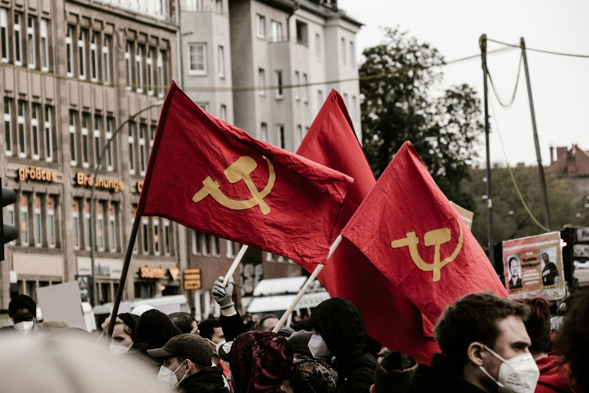 People holding red flags with yellow hammer and sickle symbols in a city street.