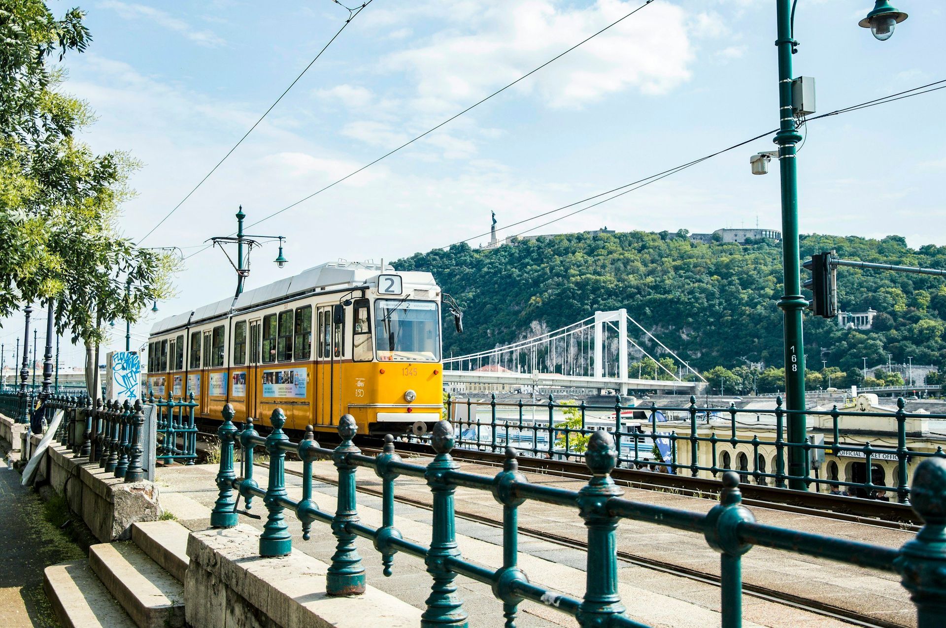 Yellow tram traveling along tracks, with a bridge and a tree-covered hill in the background. Sunny day.