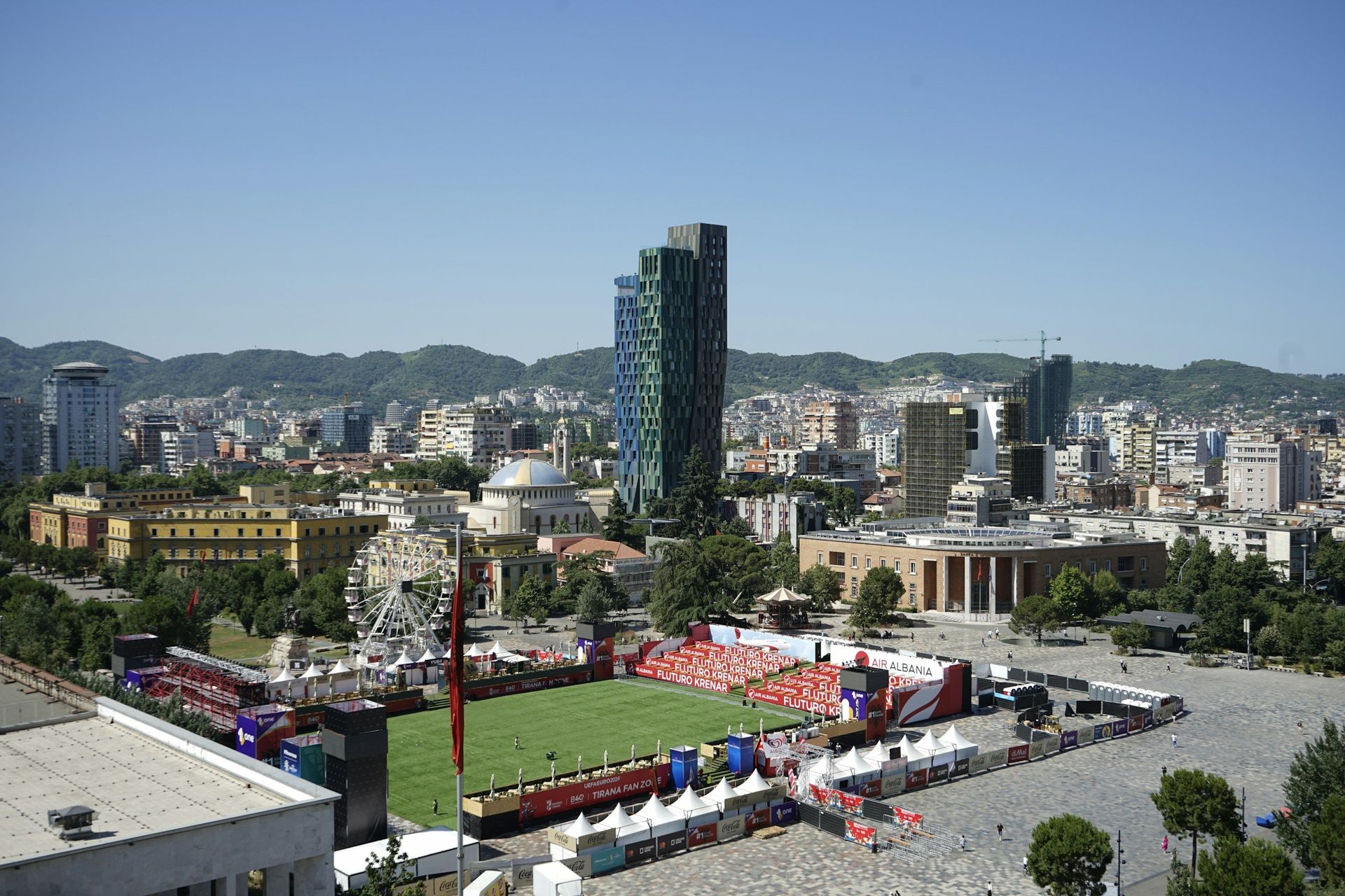 Cityscape of Tirana, Albania, with tall buildings, a Ferris wheel, and a large event in a public square under a clear blue sky.