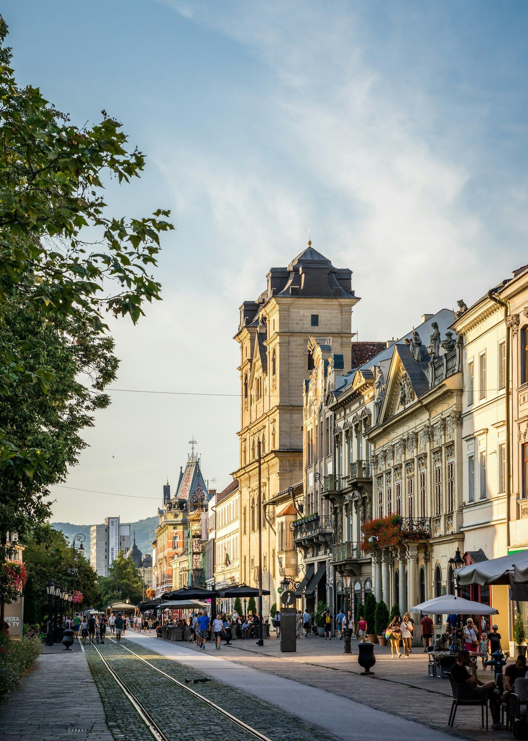 Street scene with ornate buildings, tram tracks, people, and a tall tower. Sunny, daytime.