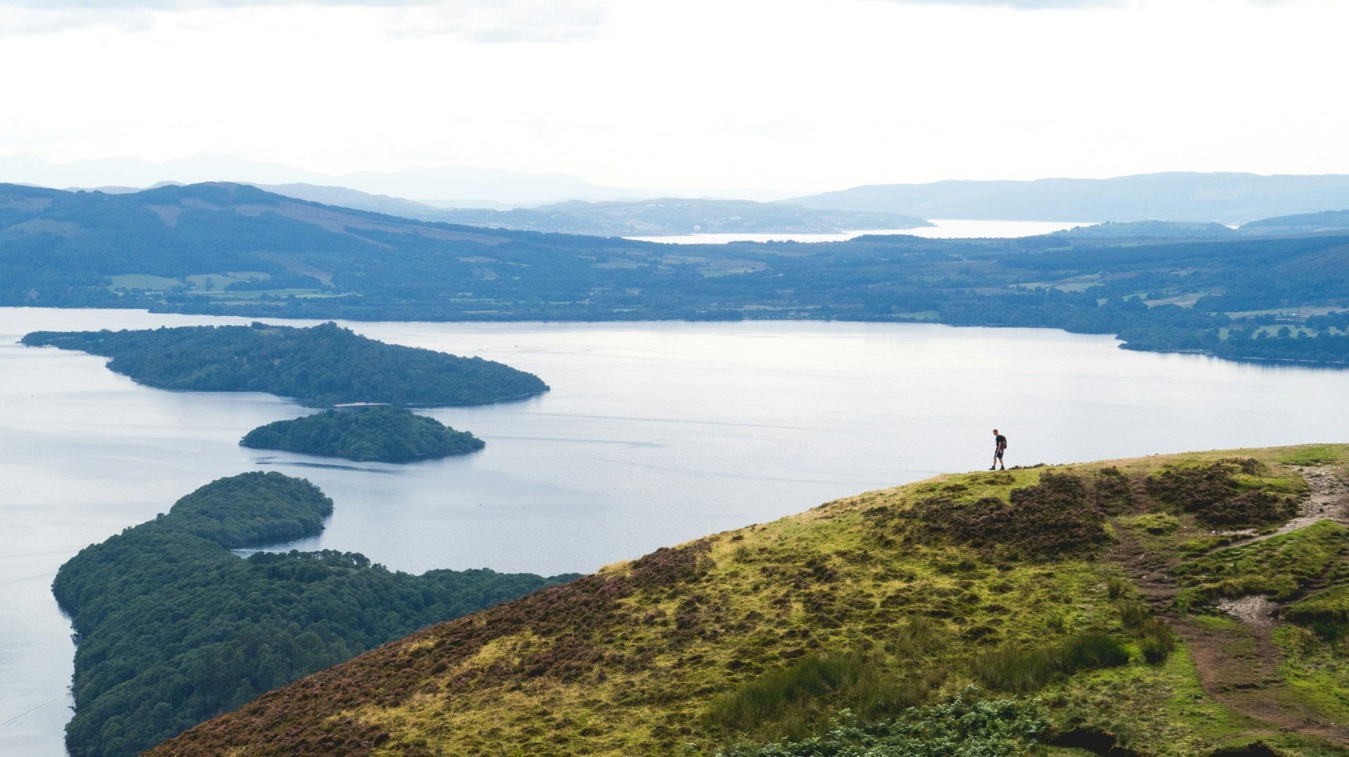 Person standing on a hillside overlooking a lake with islands and distant mountains; overcast sky.