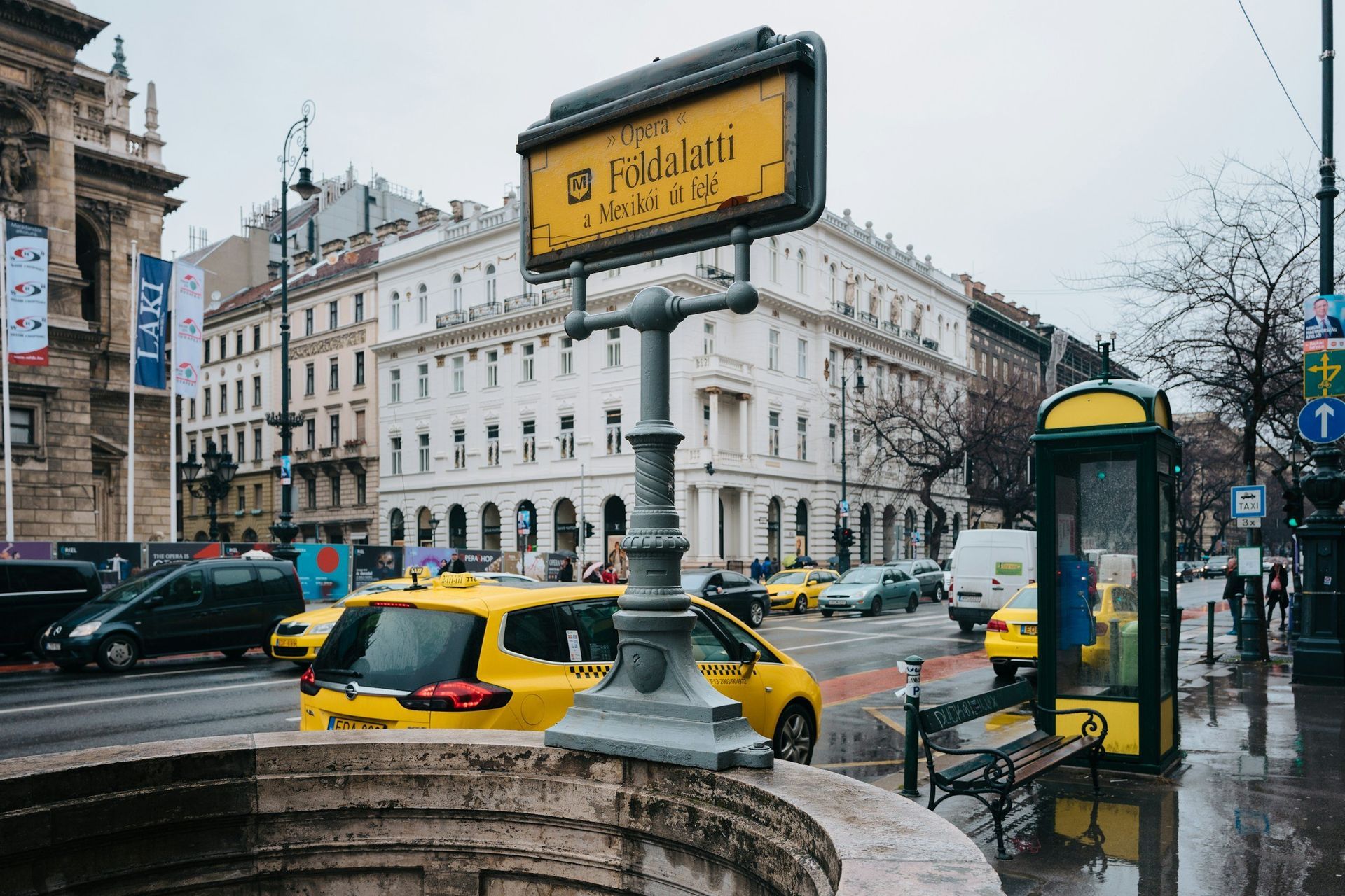 Yellow subway entrance sign in Budapest, Hungary. Yellow taxis and buildings in the background on a wet street.