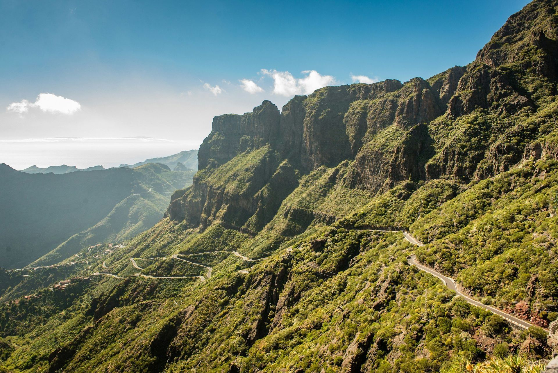 Mountainous landscape with winding road, lush green vegetation, and blue sky.