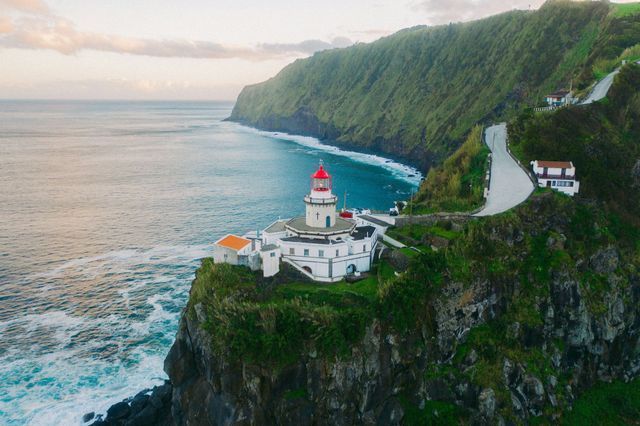 Lighthouse on a cliff overlooking the ocean, green hillside, white building with a red top, road.