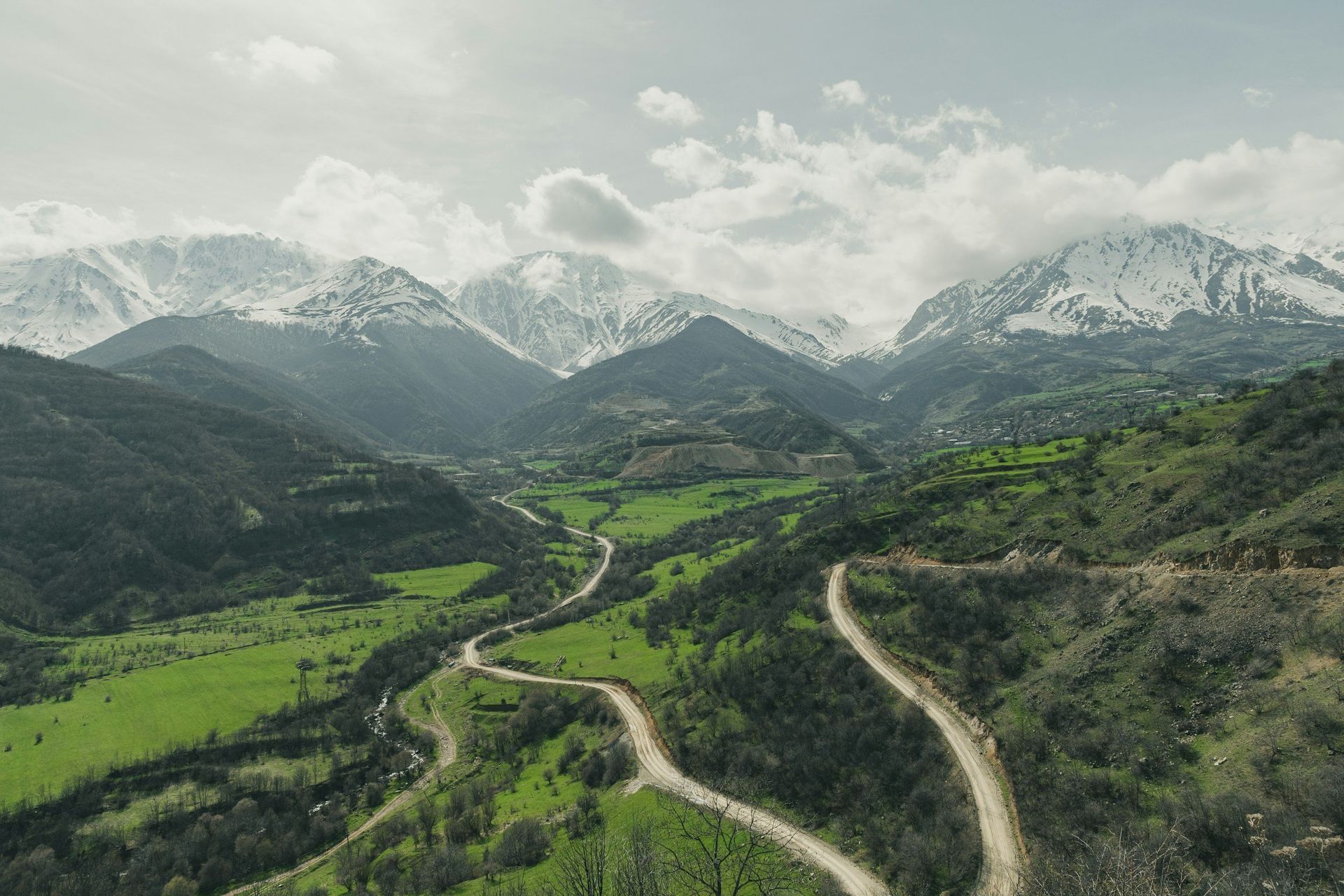 Green valley with winding road between forested slopes, snowy mountains in background.