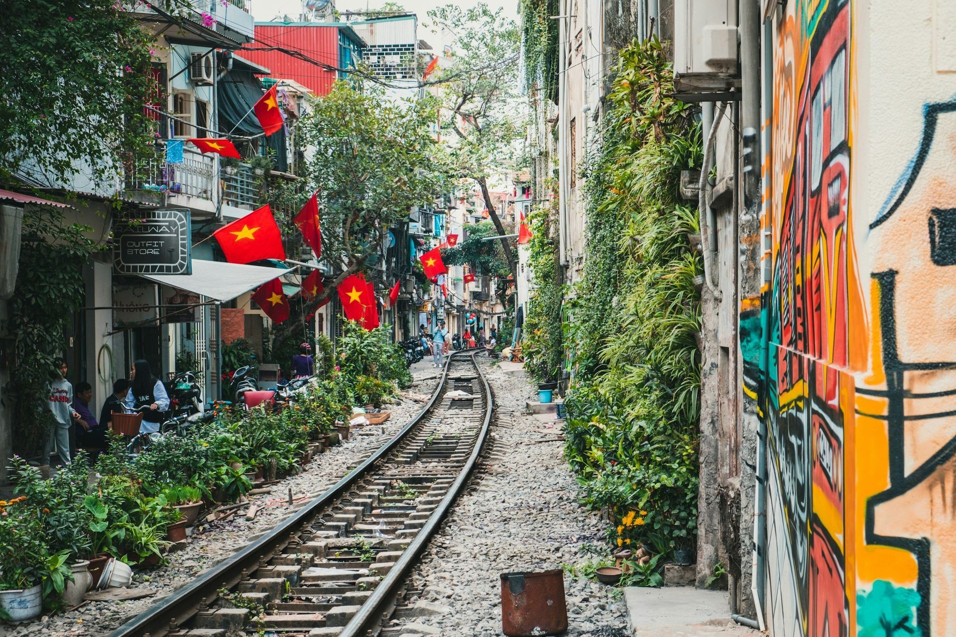 Railroad track running through a narrow alley with cafes, green plants, and Vietnamese flags. Colorful graffiti on a wall.
