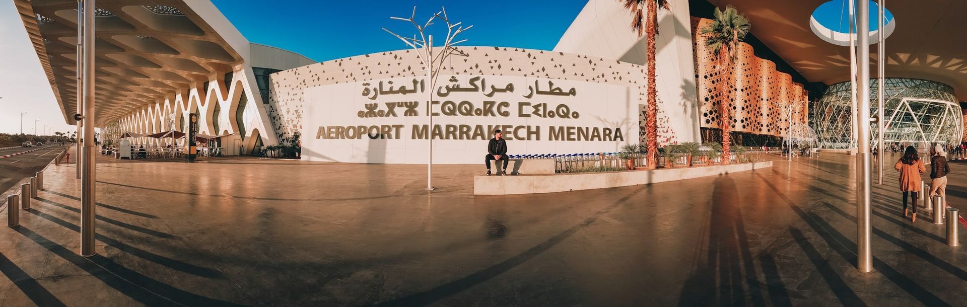 Marrakech Menara Airport exterior with Arabic and English signage. People walking near the building.