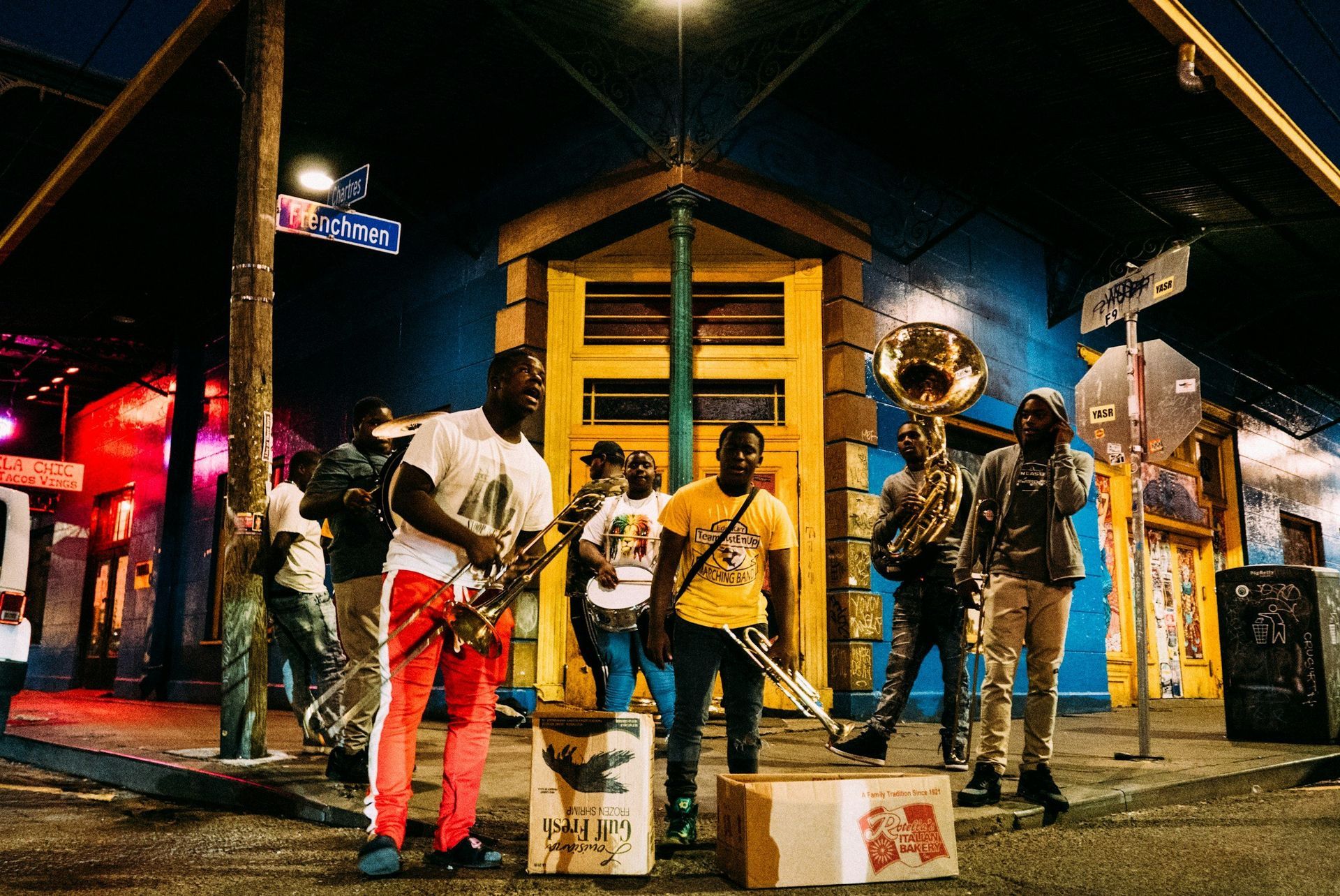 A band plays on a street at night. Musicians in casual clothes with instruments in front of a colorful building.