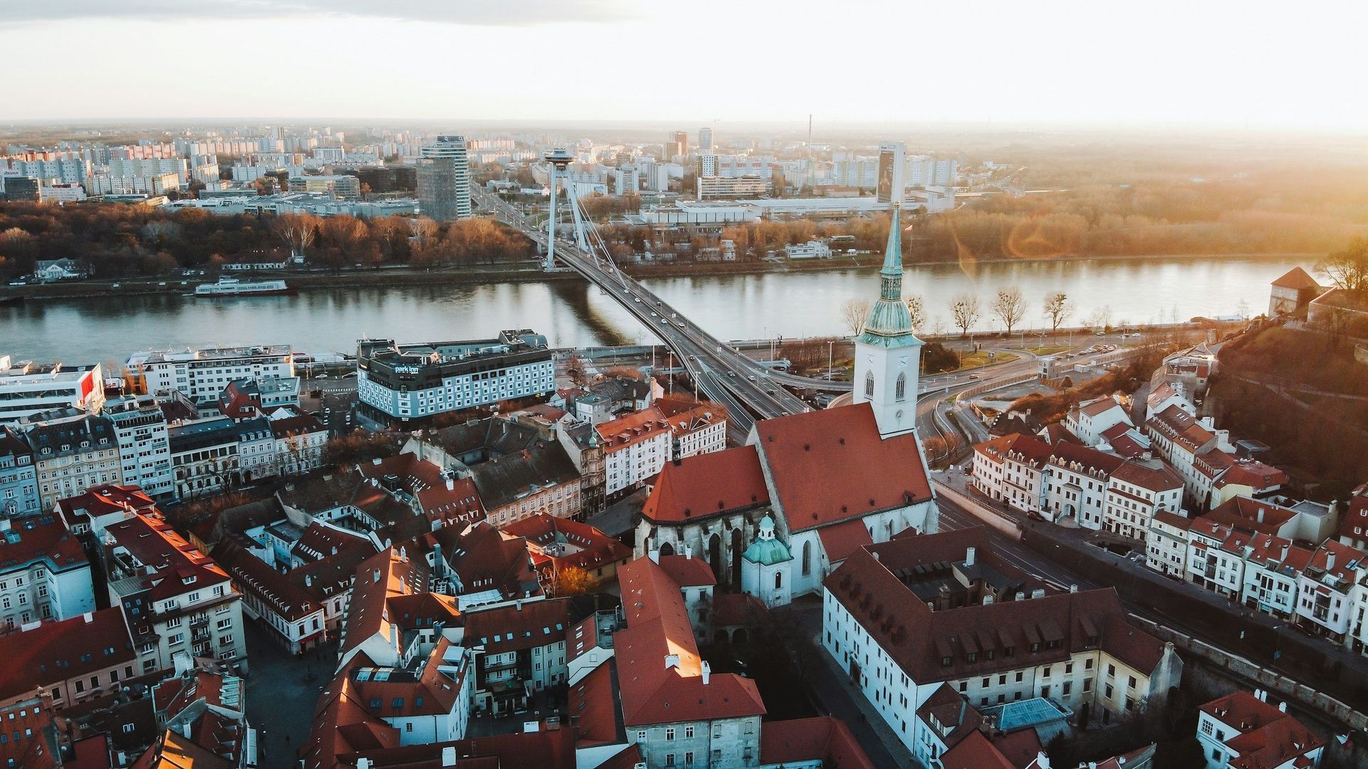 Aerial view of Bratislava, Slovakia, with St. Martin's Cathedral in foreground, bridge over river, city skyline at sunset.