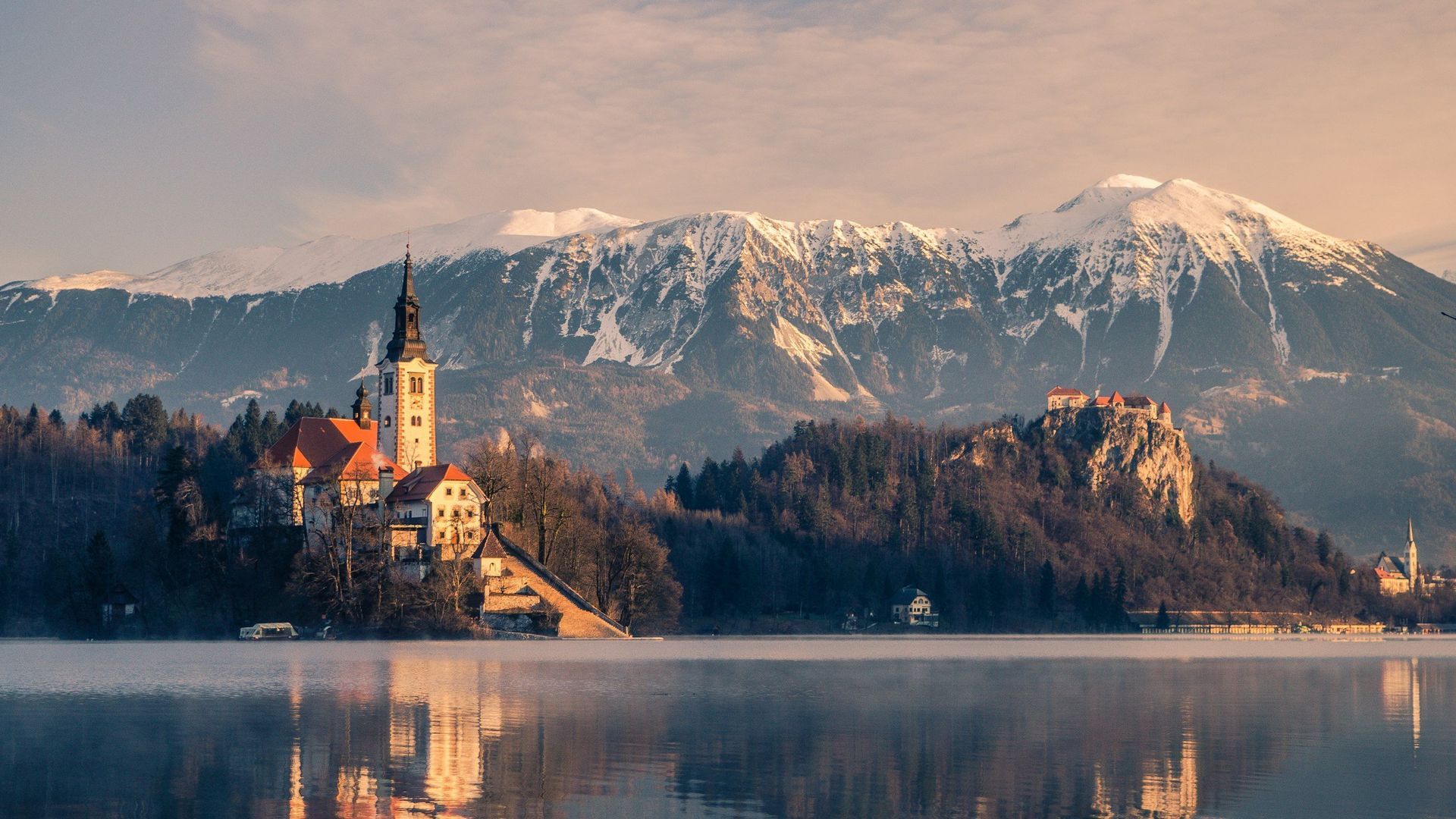 Lake Bled with church on island, castle on hill, snow-capped mountains. Still water reflects scenery.