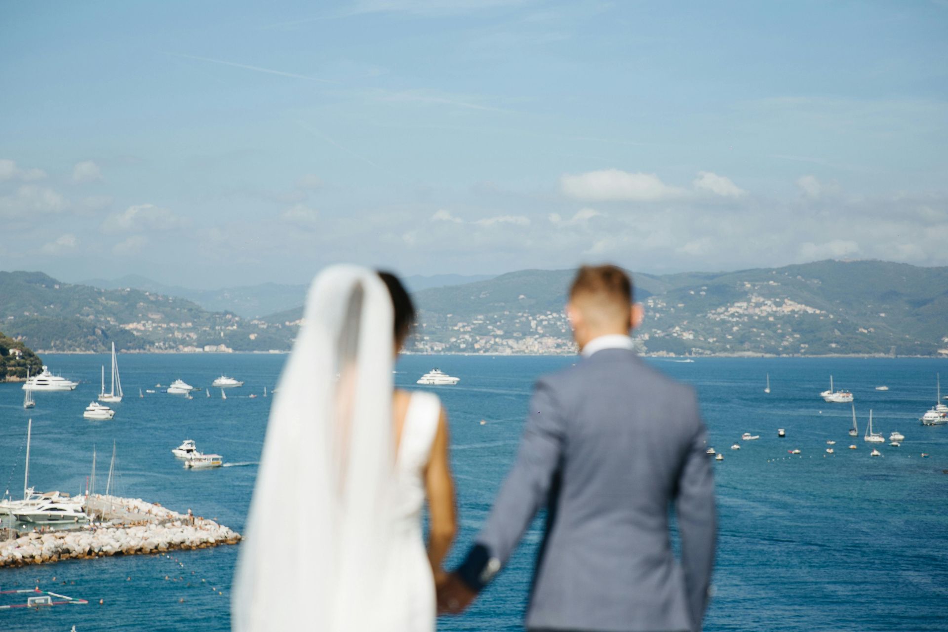 Una sposa e uno sposo si tengono per mano e guardano l'oceano.