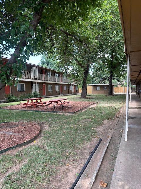 Apartment complex exterior with picnic tables, trees, and walkways. Brown and green colors dominate.