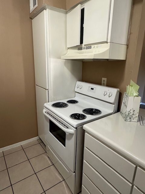 White kitchen appliances: stove, microwave, refrigerator, and cabinetry against beige walls, tiled floor.
